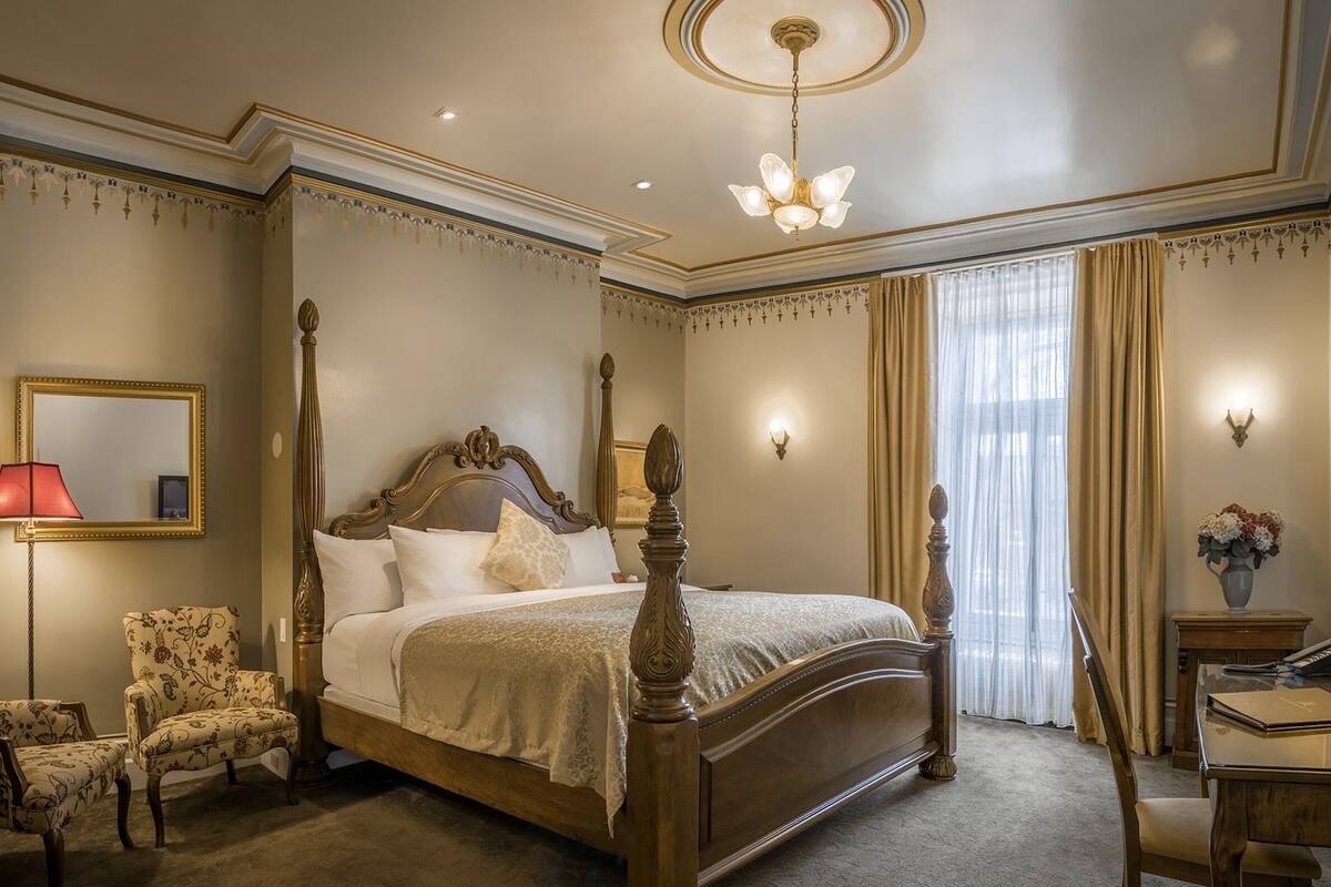 A four-poster bed with a wooden frame and a canopy, a sitting chair with a floral pattern, a desk with a vase of flowers, and a wall-mounted lamp.