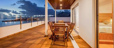Un balcón con mesa y sillas de madera, barandilla de cristal y vista a la ciudad al atardecer.