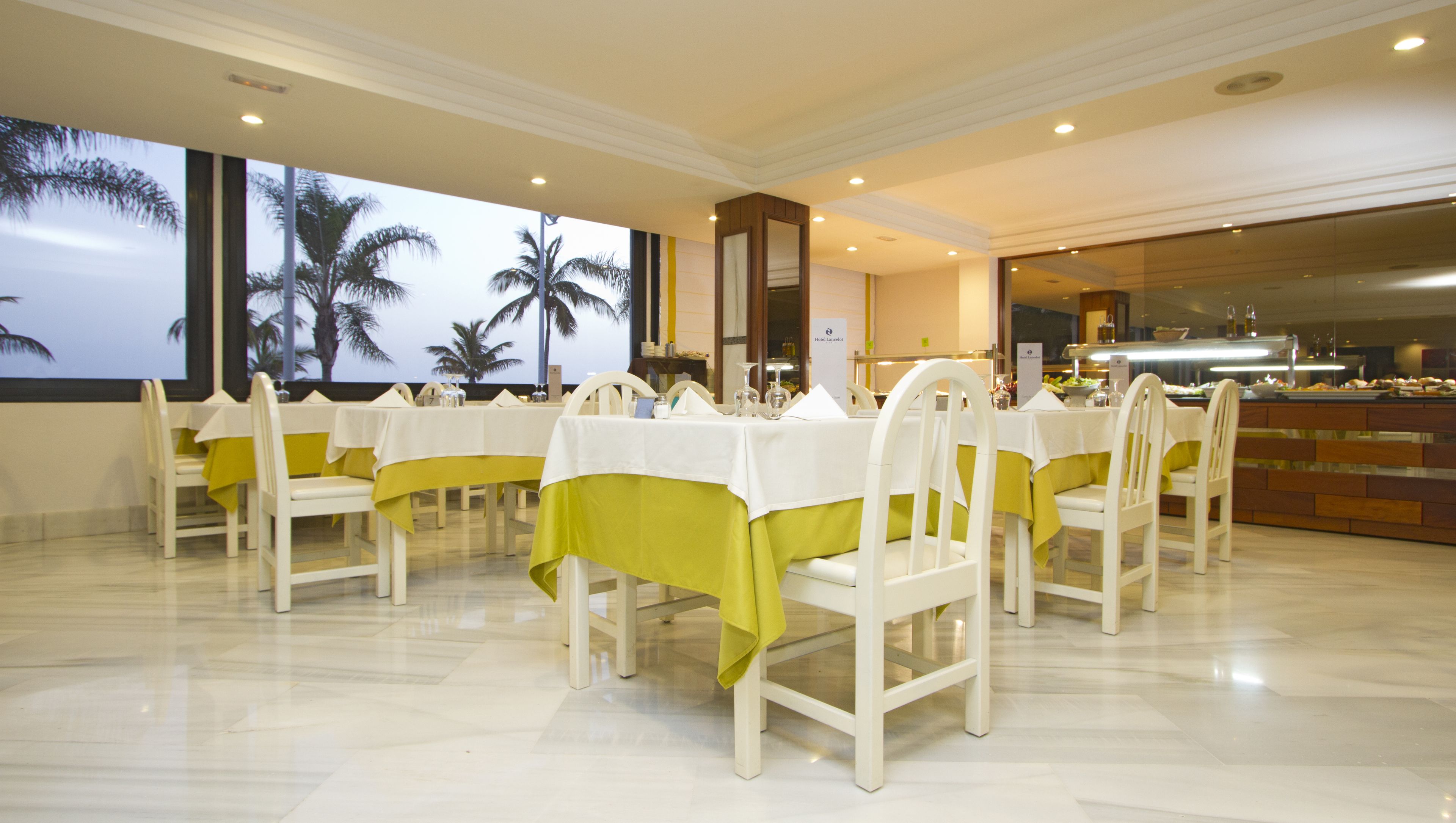 A hotel dining area with white chairs and tables, a buffet counter, and a large window with a view of palm trees.