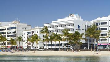 Un hotel frente a la playa con varios pisos, palmeras y una playa de arena.