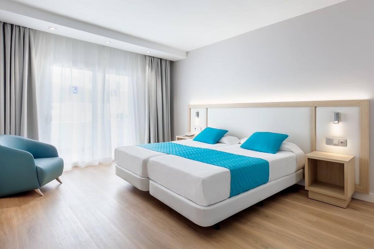 Una habitación de hotel moderna con una cama doble, una mesita de noche, una silla y un ventanal con cortinas.