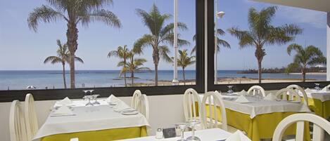 A restaurant with tables set for dining, overlooking a beach with palm trees and the ocean.