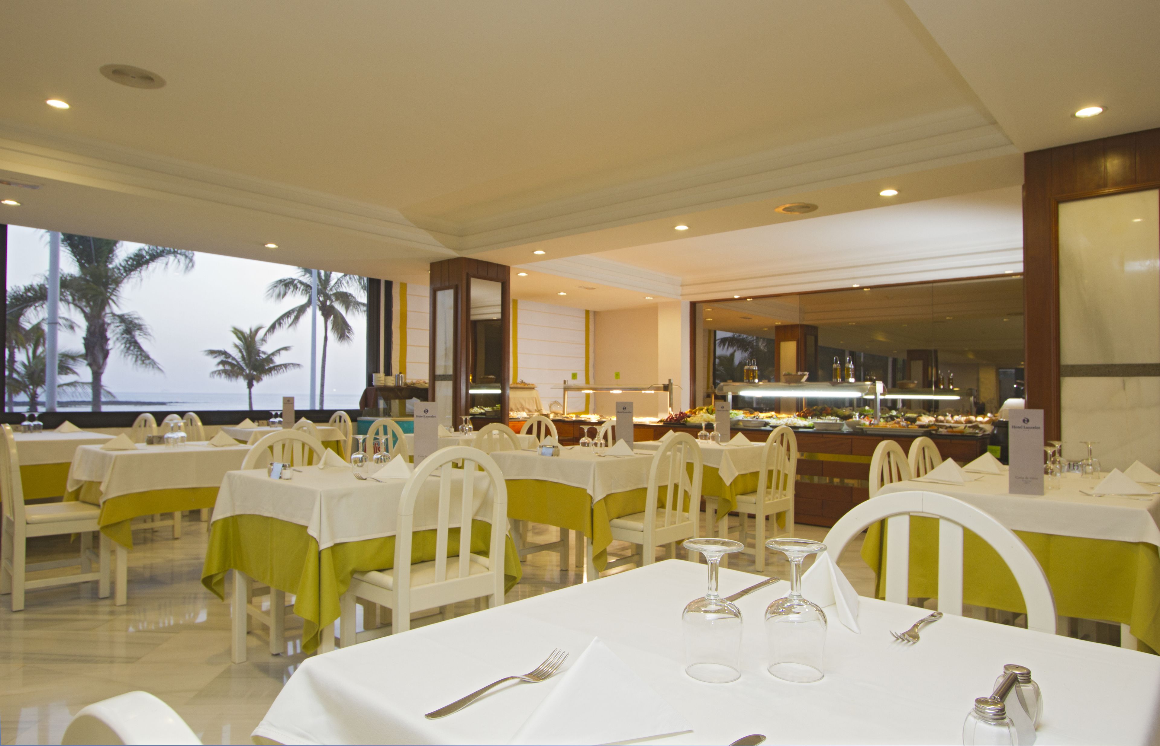 A restaurant with white tables and chairs, a buffet area, and a view of palm trees and the ocean.