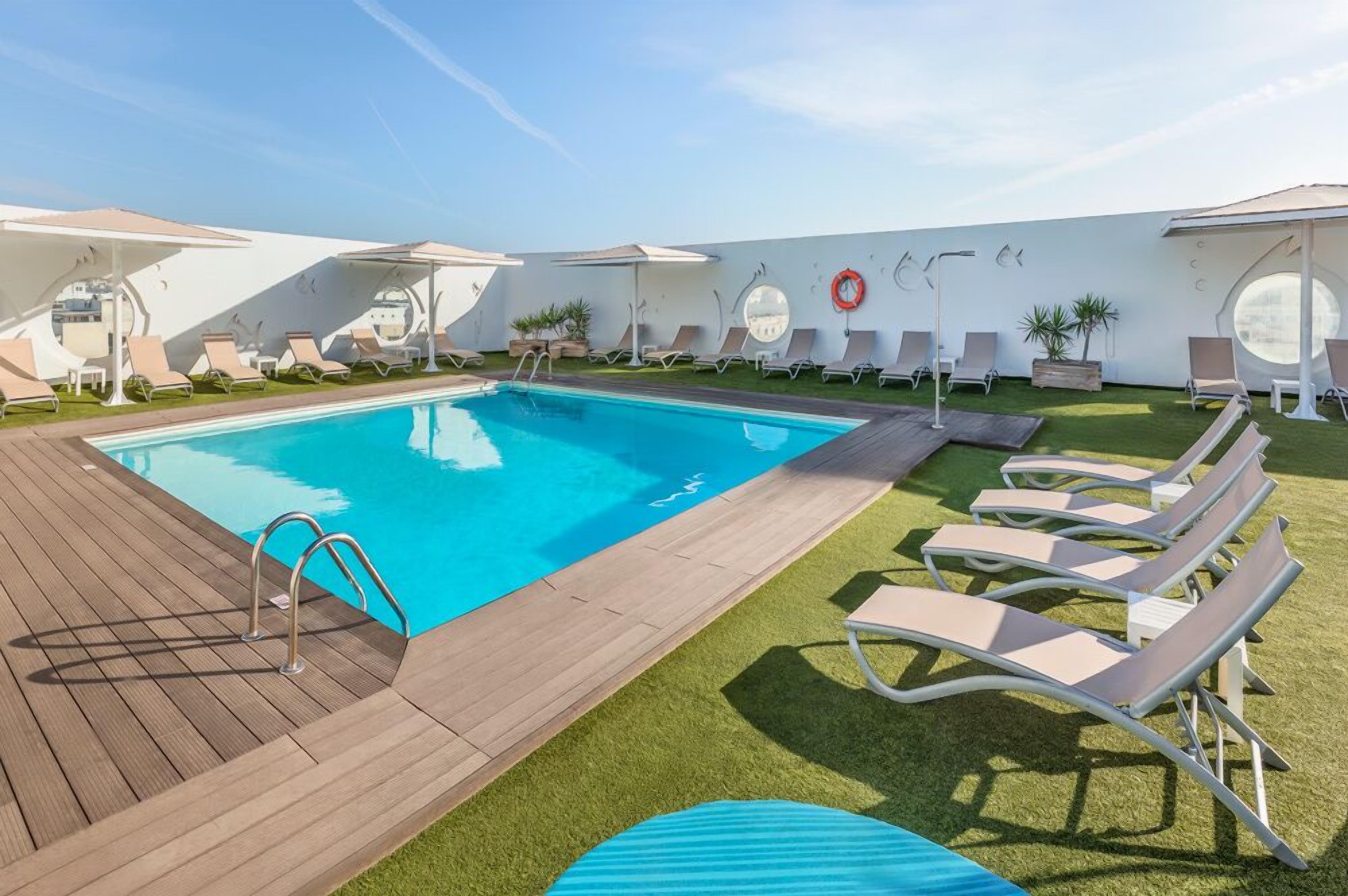 A swimming pool with a wooden deck, surrounded by lounge chairs and umbrellas.