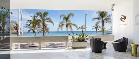 Ett modernt hotellrum med ett stort fönster med utsikt över en strand, inrett med en bänk, flätade stolar och en krukväxt.