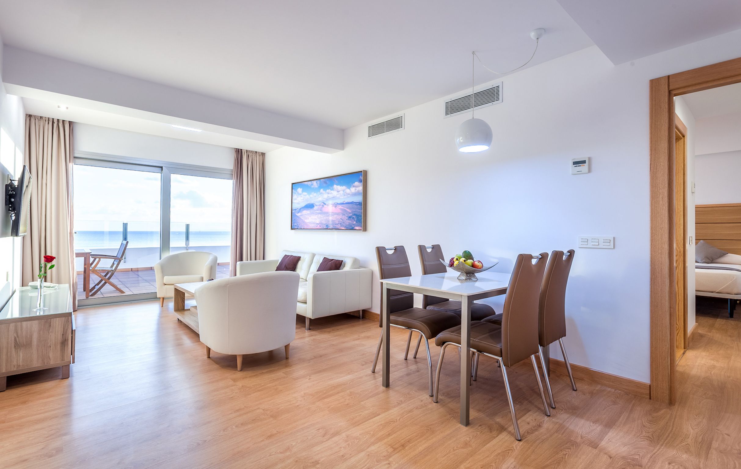 Habitación de hotel moderna con zona de comedor, sofá, mesa y sillas, y un gran ventanal con vistas al mar.
