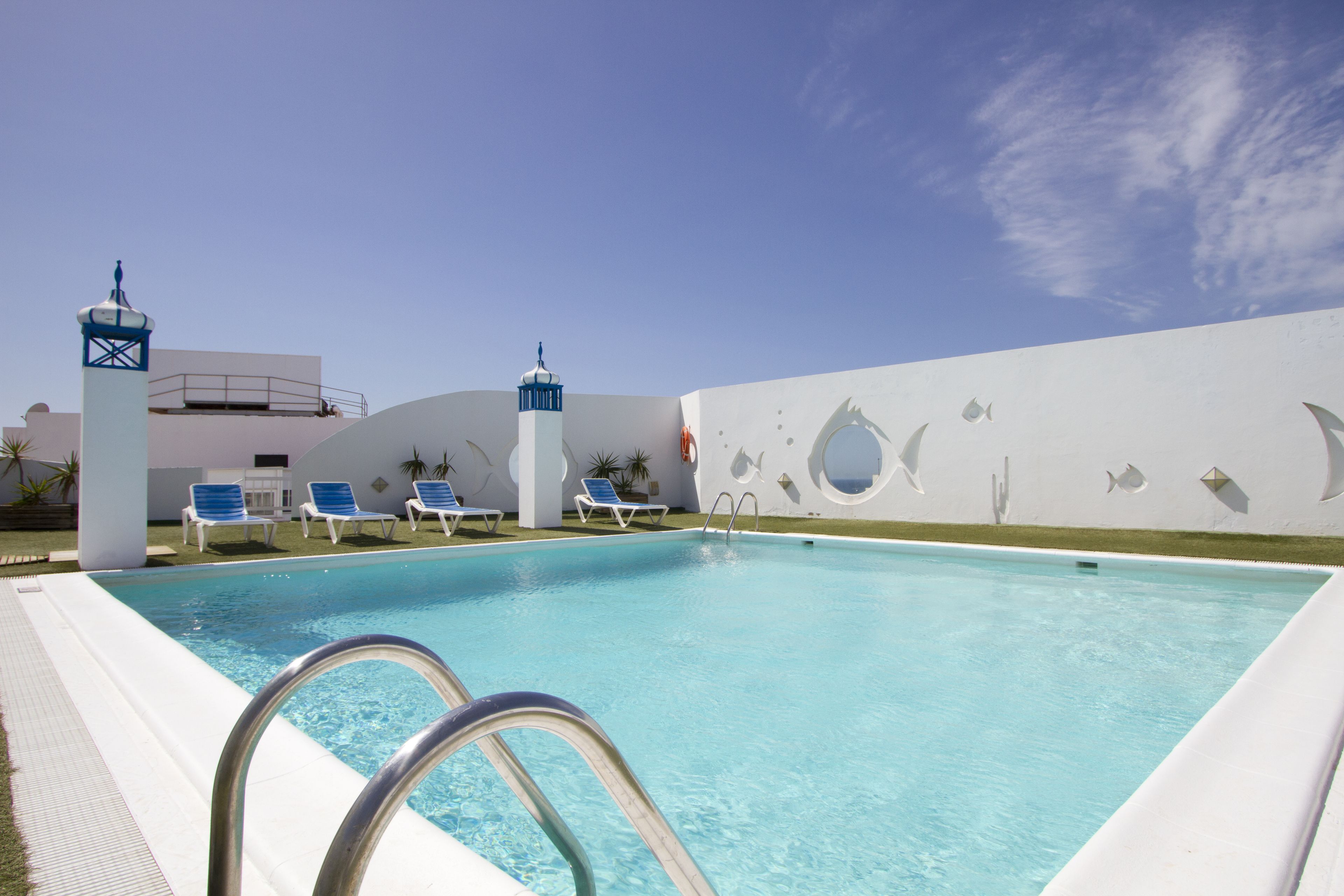 Una piscina con agua cristalina azul, rodeada de sillas de descanso y un muro decorativo con motivos de peces.