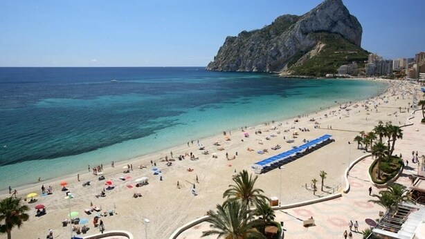 Uma praia com águas turquesa cristalinas, uma colina rochosa ao fundo e um calçadão com palmeiras.