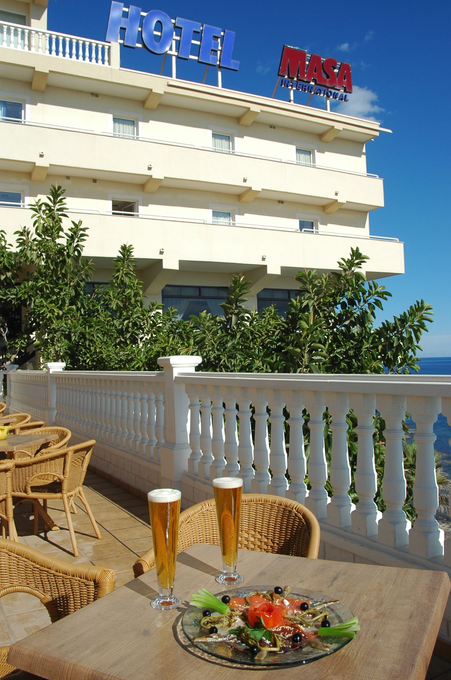 A hotel with a terrace featuring wicker furniture and a table with two glasses of beer and a plate of salad.