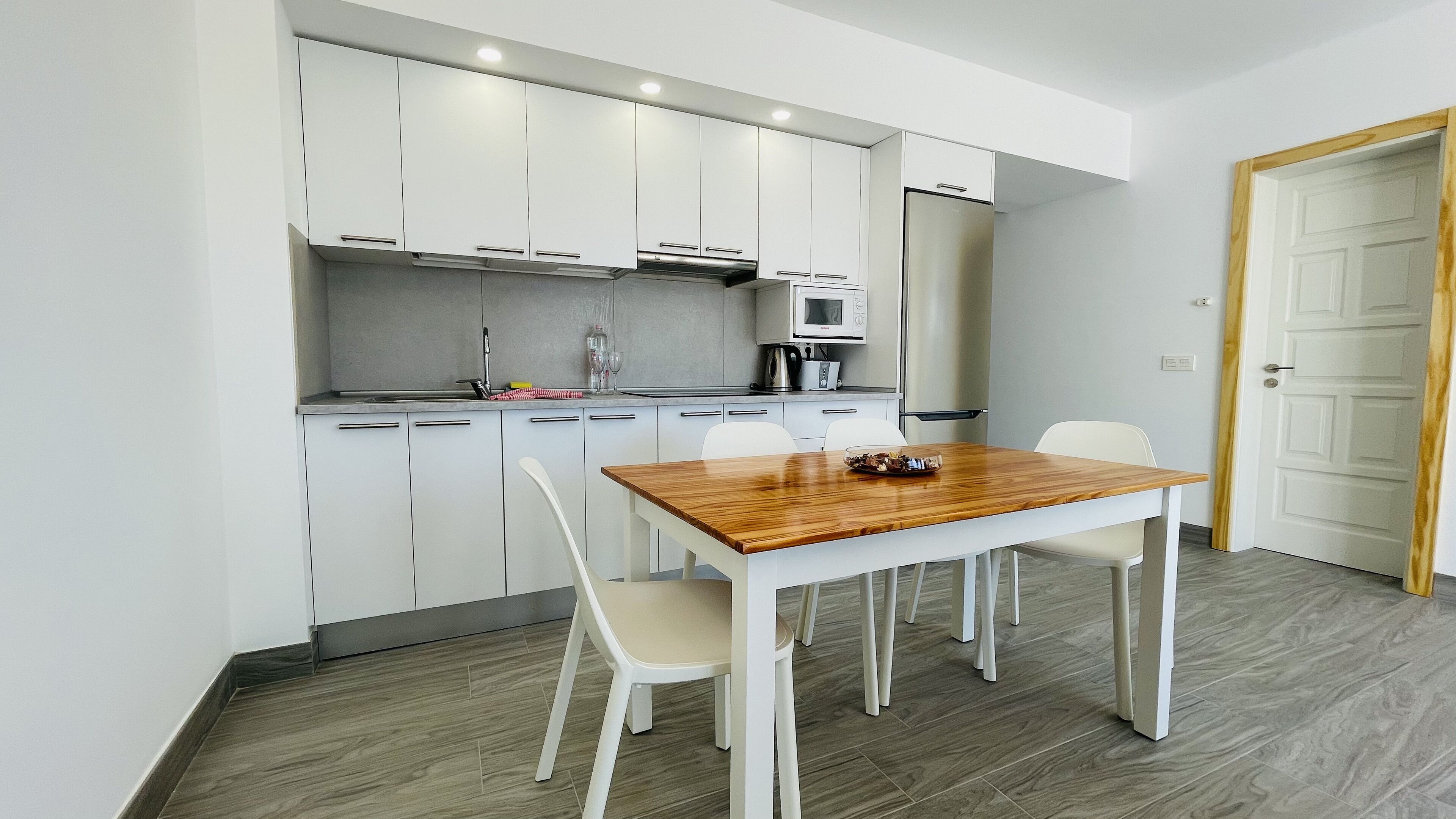 Een moderne keuken met witte kasten, een houten eettafel en een witte deur.