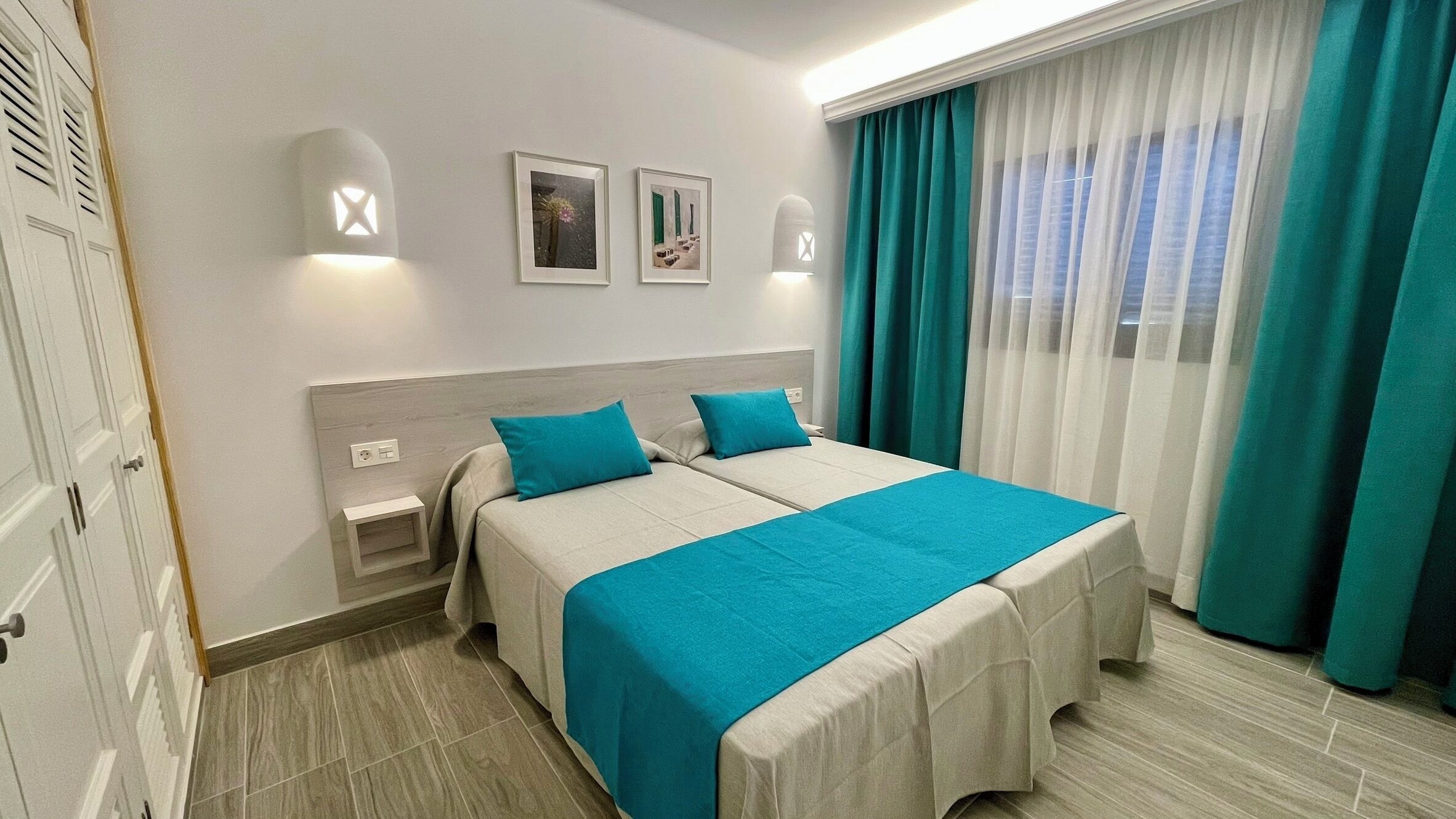 Une chambre d’hôtel avec deux lits, une table de chevet, une lampe fixée au mur et une fenêtre avec des rideaux.