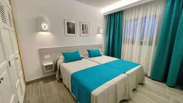 Een hotelkamer met twee bedden, een nachtkastje, een aan de muur bevestigde lamp en een raam met gordijnen.