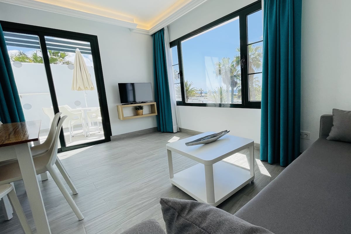 Ein modernes Wohnzimmer mit Sofa, Fernseher auf einem Regal, Essbereich mit Stühlen und großen Fenstern mit Vorhängen.