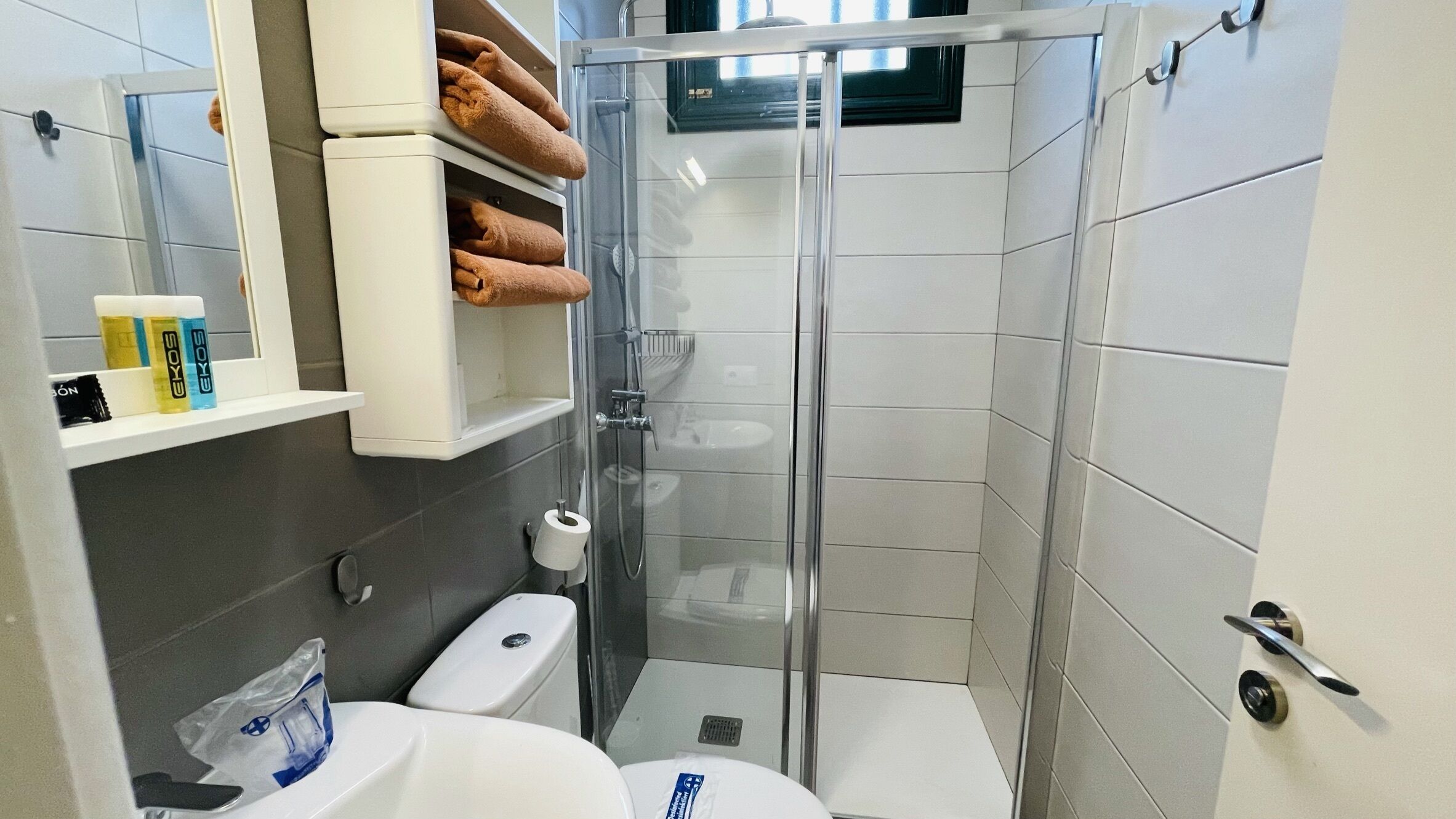 A modern bathroom with a glass-enclosed shower, a toilet, and a sink. There are towels and toiletries on a shelf.