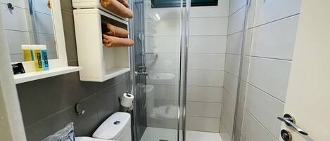A modern bathroom with a glass-enclosed shower, a toilet, and a sink. There are towels and toiletries on a shelf.