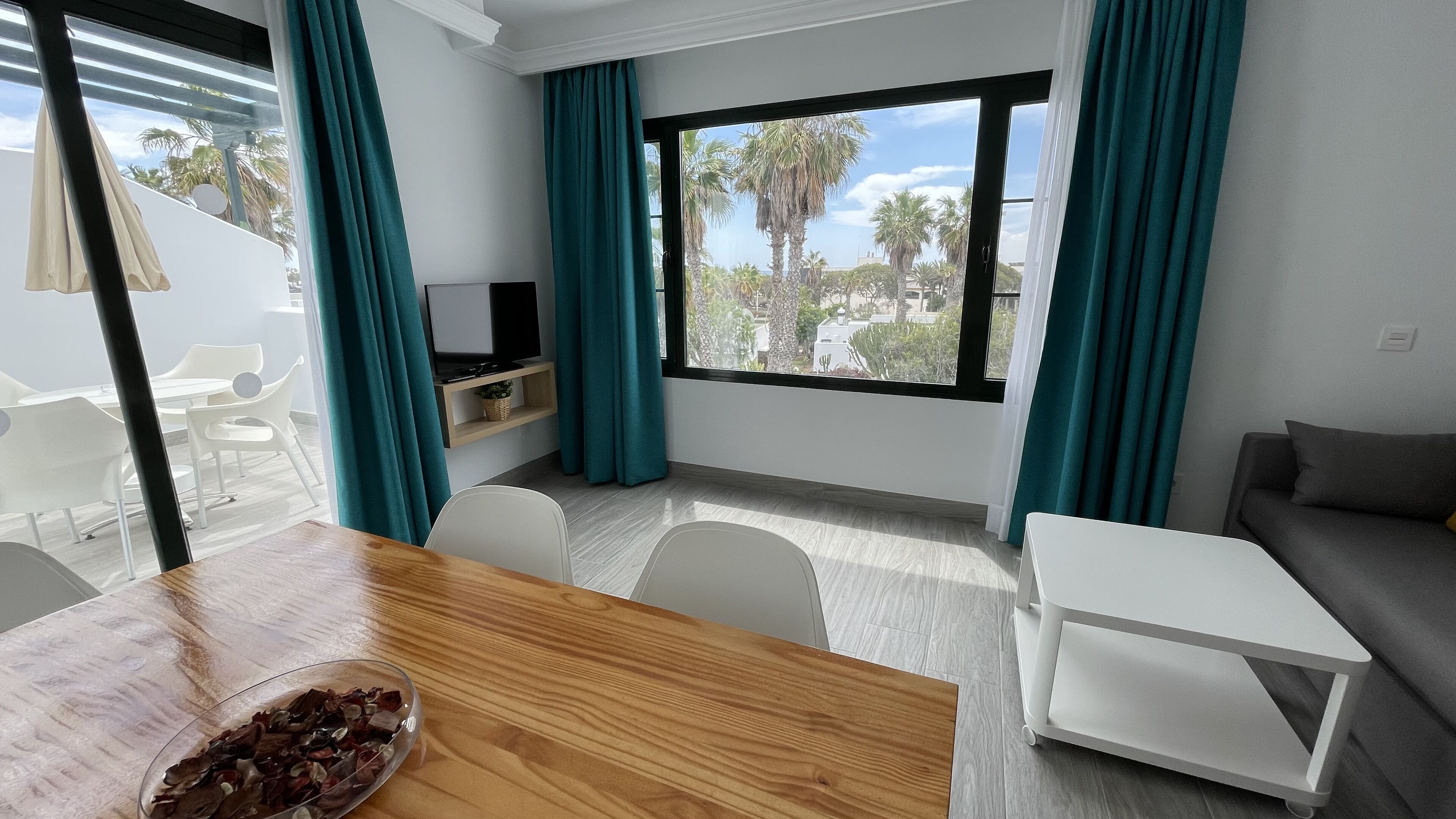 Ein modernes Wohnzimmer mit einem Holzesstisch, einem grauen Sofa und einem großen Fenster mit türkisfarbenen Vorhängen.