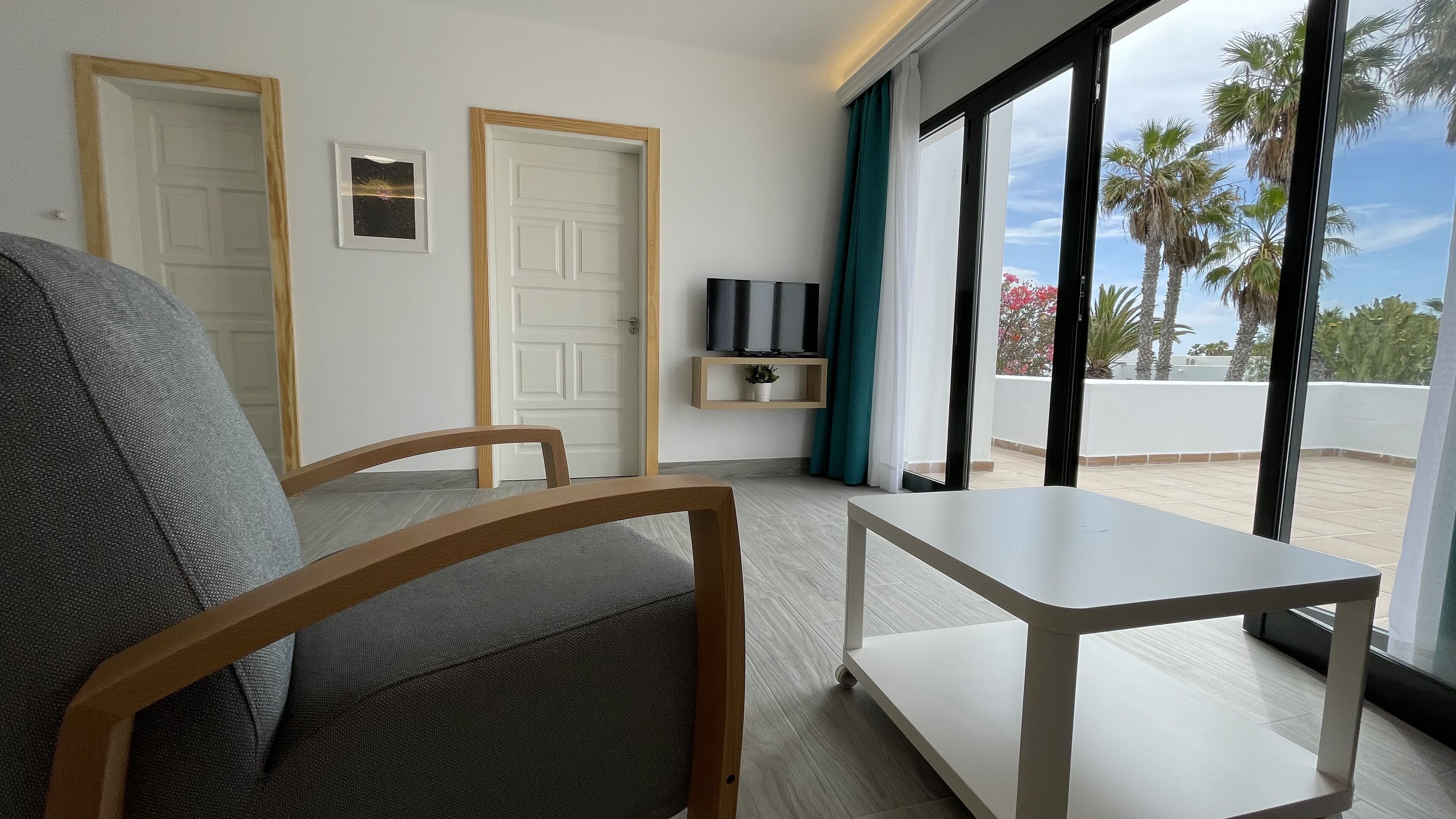 A room with a sofa, a small table, a TV on a shelf, and a view of palm trees through large windows.