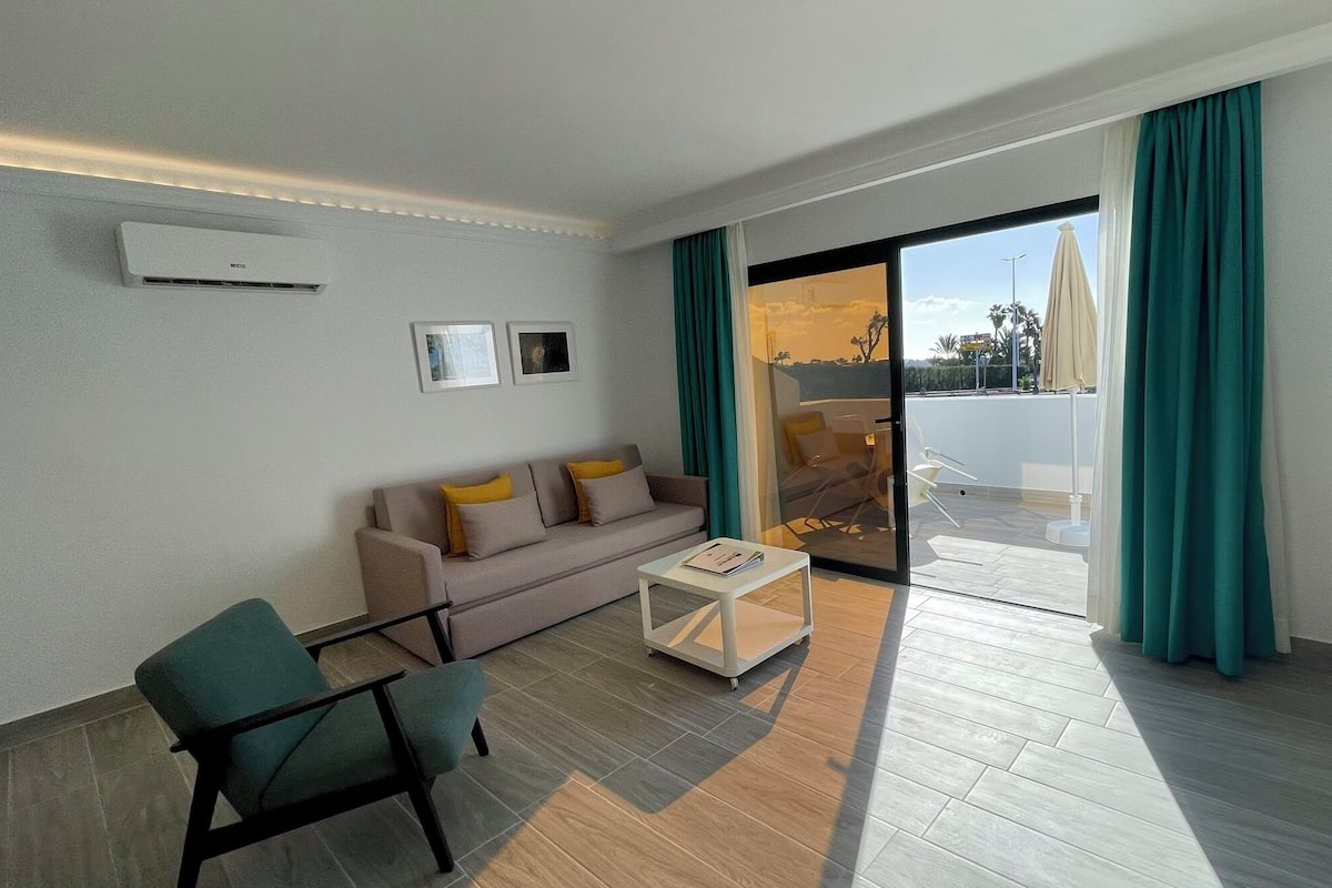 Ein modernes Wohnzimmer mit einer Couch, einem Sessel und einem Couchtisch. Eine Glastür führt zu einem Außenbereich mit Pool und Sitzgelegenheiten.