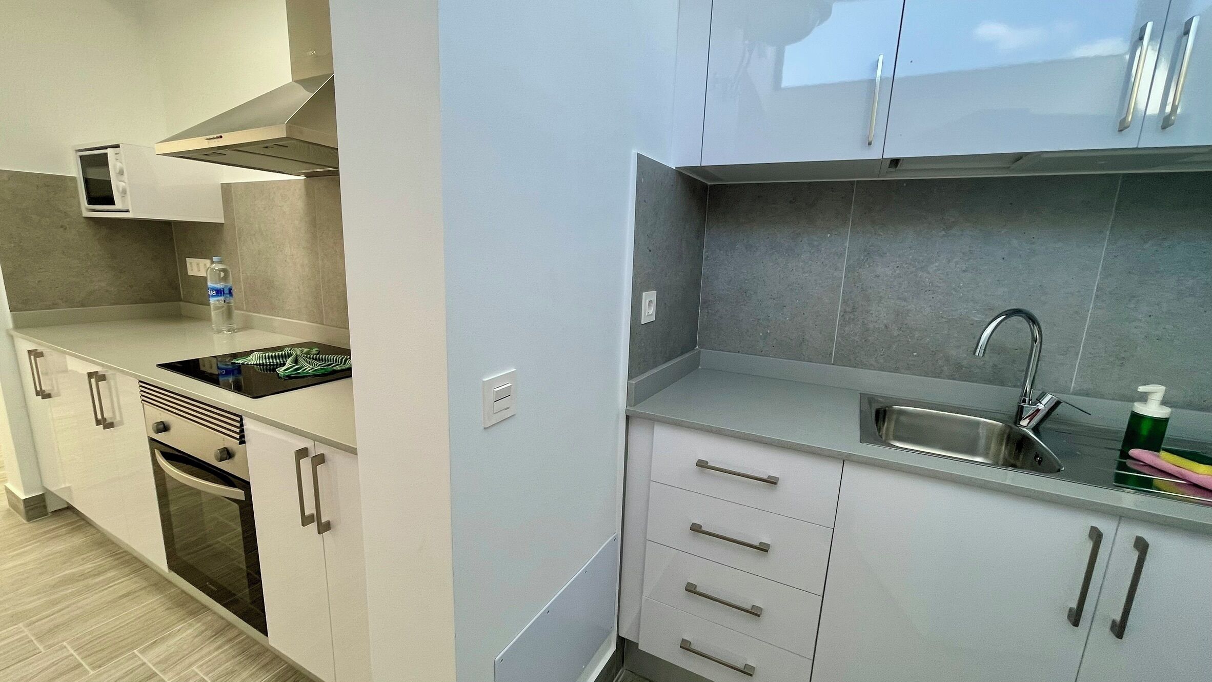Una cocina moderna con microondas integrado, estufa y fregadero. Hay armarios con tiradores y una encimera con una botella de detergente.