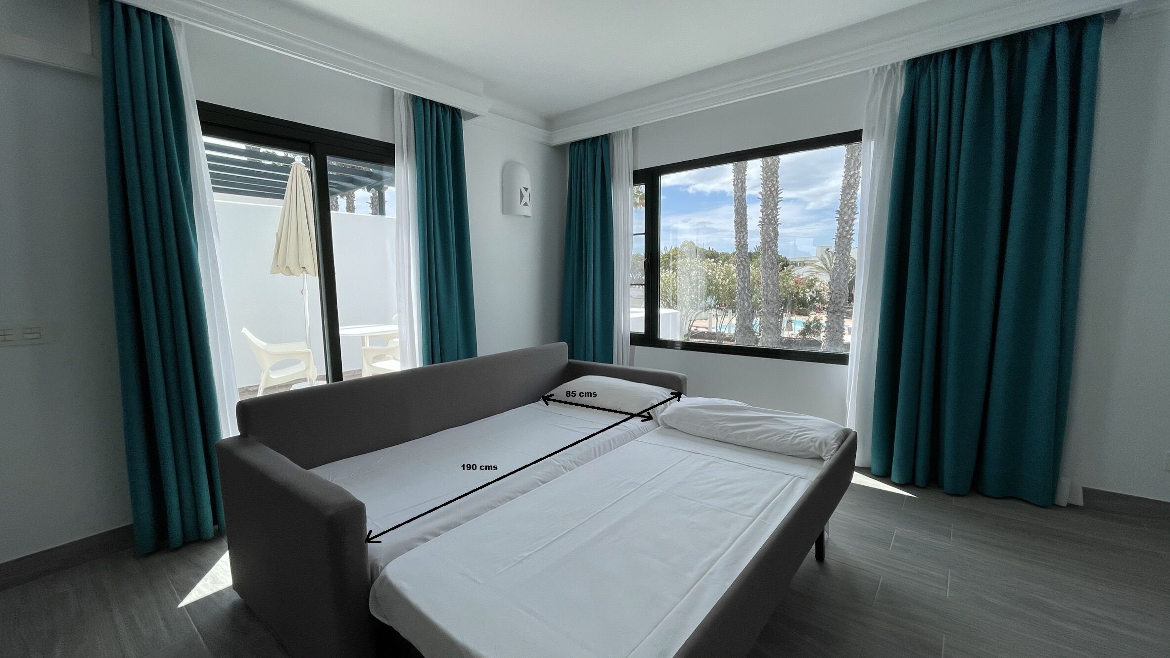 A modern hotel room with a bed measuring 190 cms in length and 85 cms in width, large windows with teal curtains, and a view of palm trees and a pool.