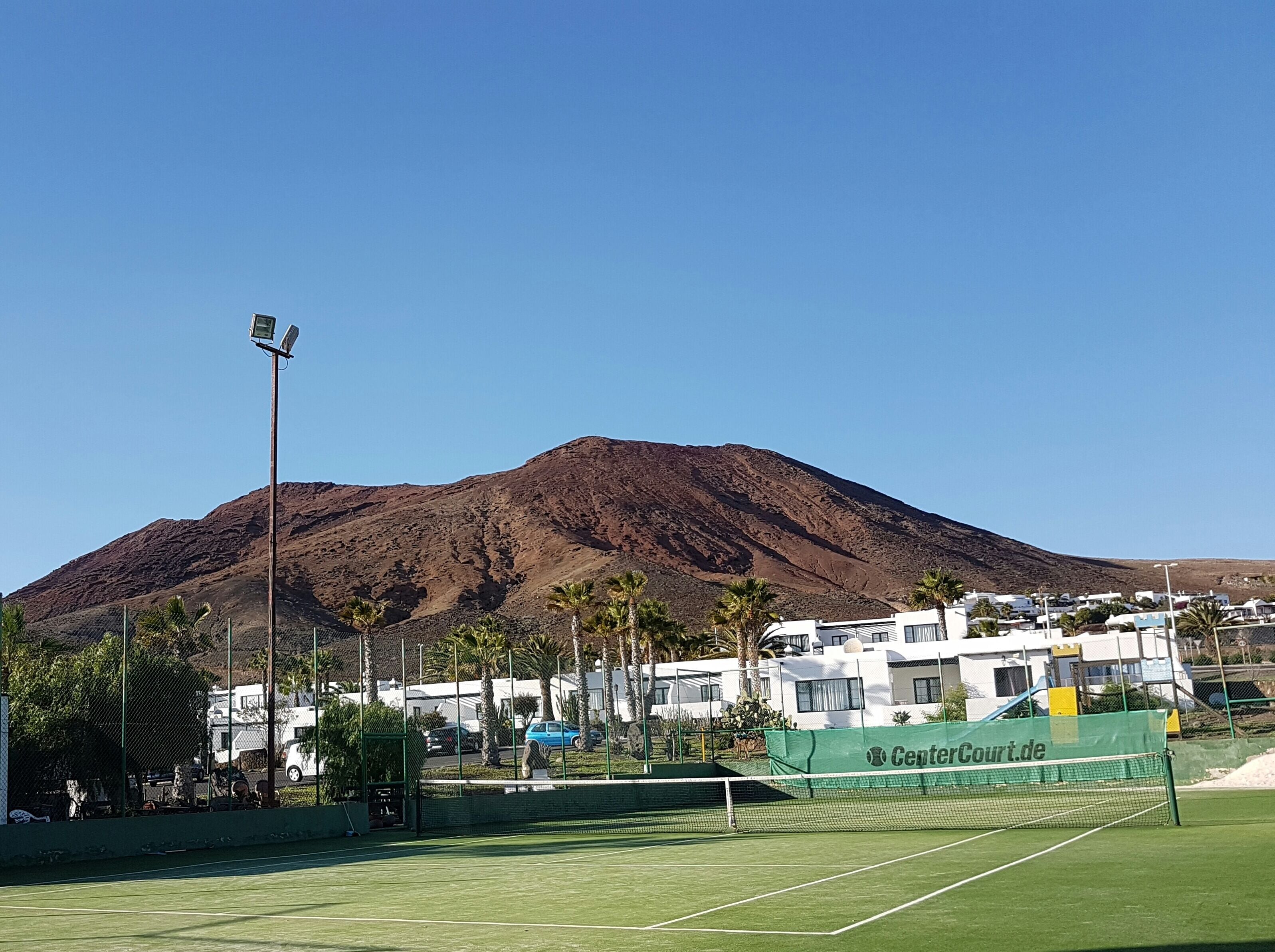Un court de tennis avec un filet, entouré d’une clôture, et une montagne en arrière-plan.