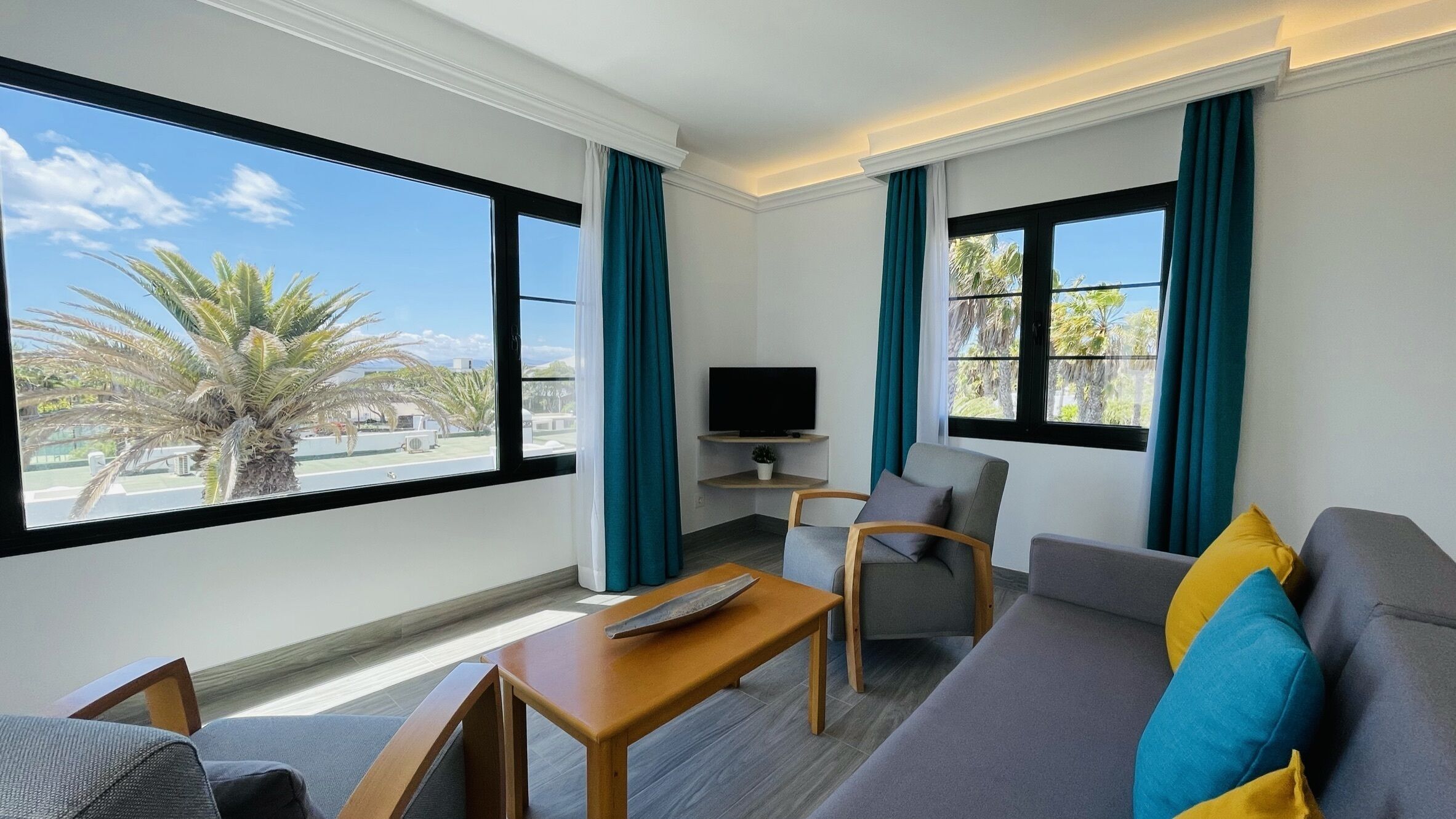 A modern hotel room with a sofa, a small table, a TV, and large windows offering a view of palm trees.