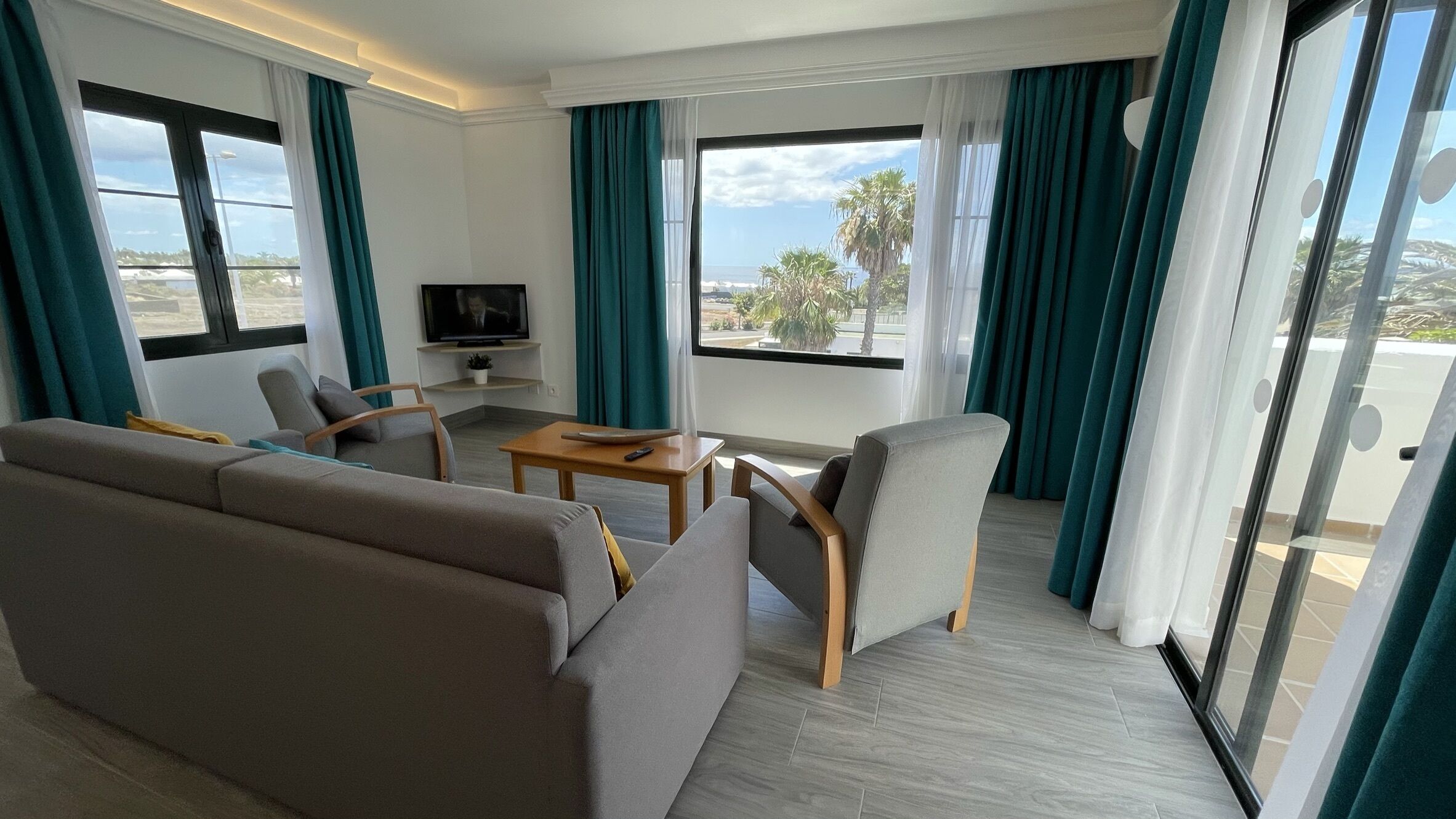 Una sala de estar moderna con un sofá, sillones, una mesa de centro y un televisor. Amplias ventanas con cortinas ofrecen vistas a palmeras y al océano.