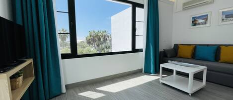 Ein modernes Wohnzimmer mit einem großen Fenster, einem Sofa mit bunten Kissen, einem weißen Couchtisch und einem Fernseher auf einem Holzbodenregal.