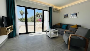 A modern living room with a grey sofa, a coffee table, and a TV on a wooden shelf. There is a sliding glass door leading to an outdoor area with a patio and palm trees. - Palmeras Garden (Yaiza)