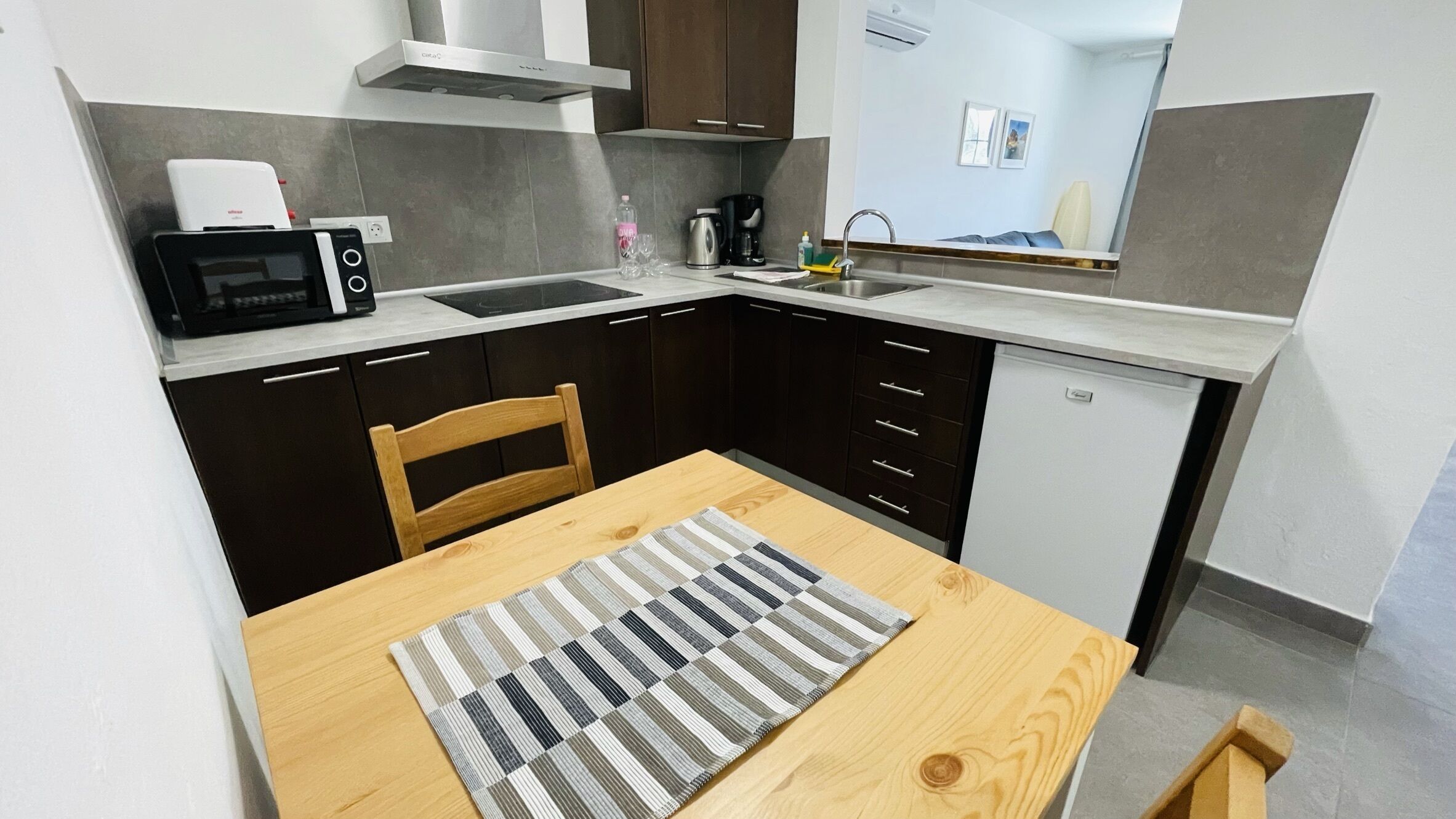 A compact kitchen with dark cabinets, a microwave, a coffee maker, and a sink. There's a wooden table with a striped placemat.