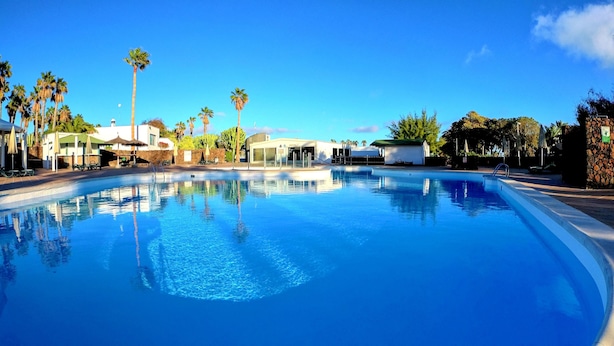 A clear blue swimming pool surrounded by palm trees and a white building with a green roof. - Palmeras Garden (Yaiza)