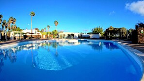 A clear blue swimming pool surrounded by palm trees and a white building with a green roof. - Palmeras Garden (Yaiza)