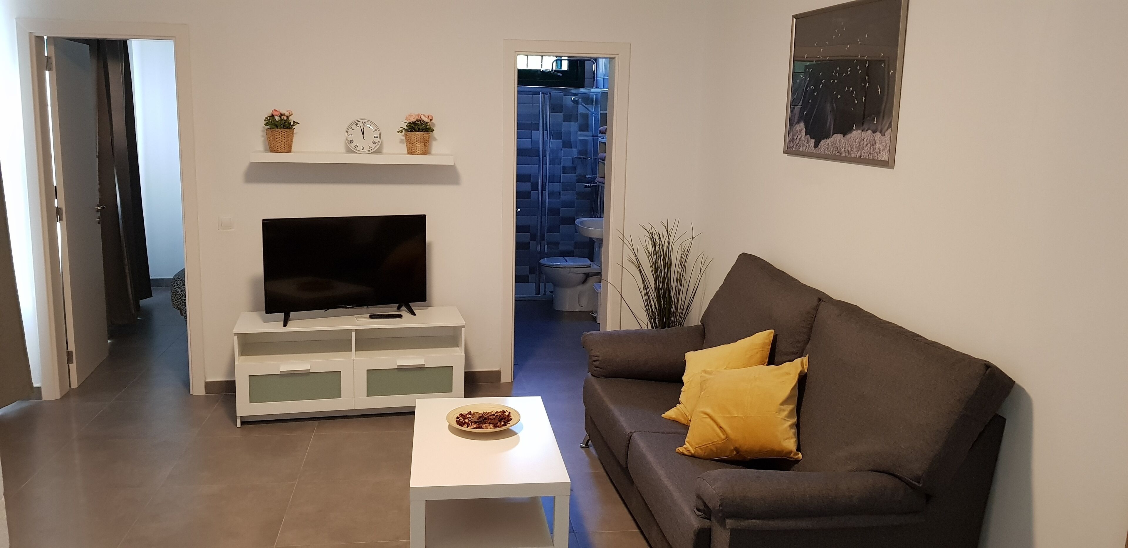 Ein Wohnzimmer mit einem Flachbildfernseher auf einem weißen Standfuß, einem braunen Sofa mit einem gelben Kissen, einem kleinen weißen Tisch mit einer Schüssel und einer Wandregal mit zwei Uhren und zwei Blumentöpfen.
