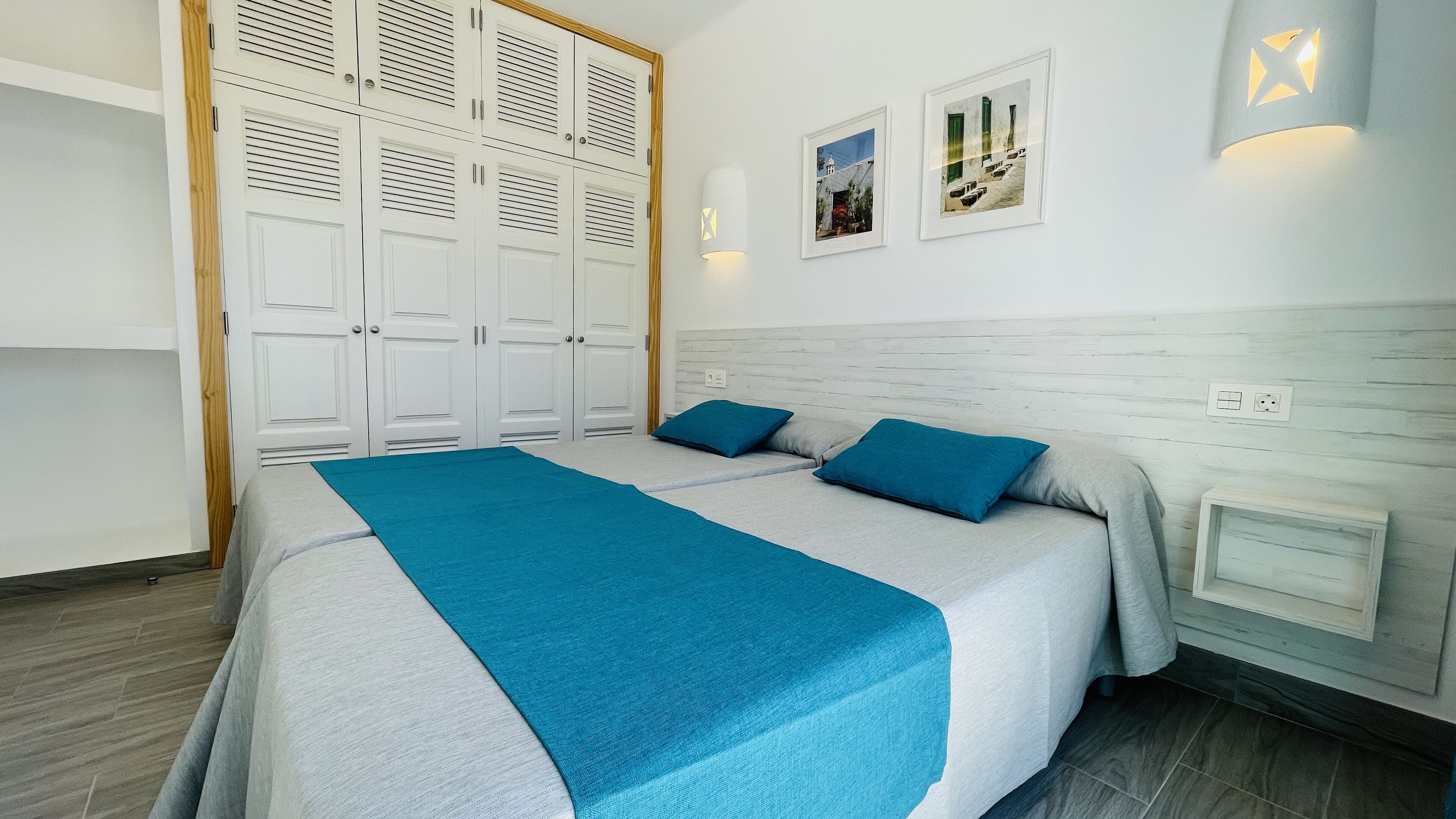 A bedroom with a bed, two pillows, a nightstand, a wall-mounted lamp, and a wardrobe with louvered doors.