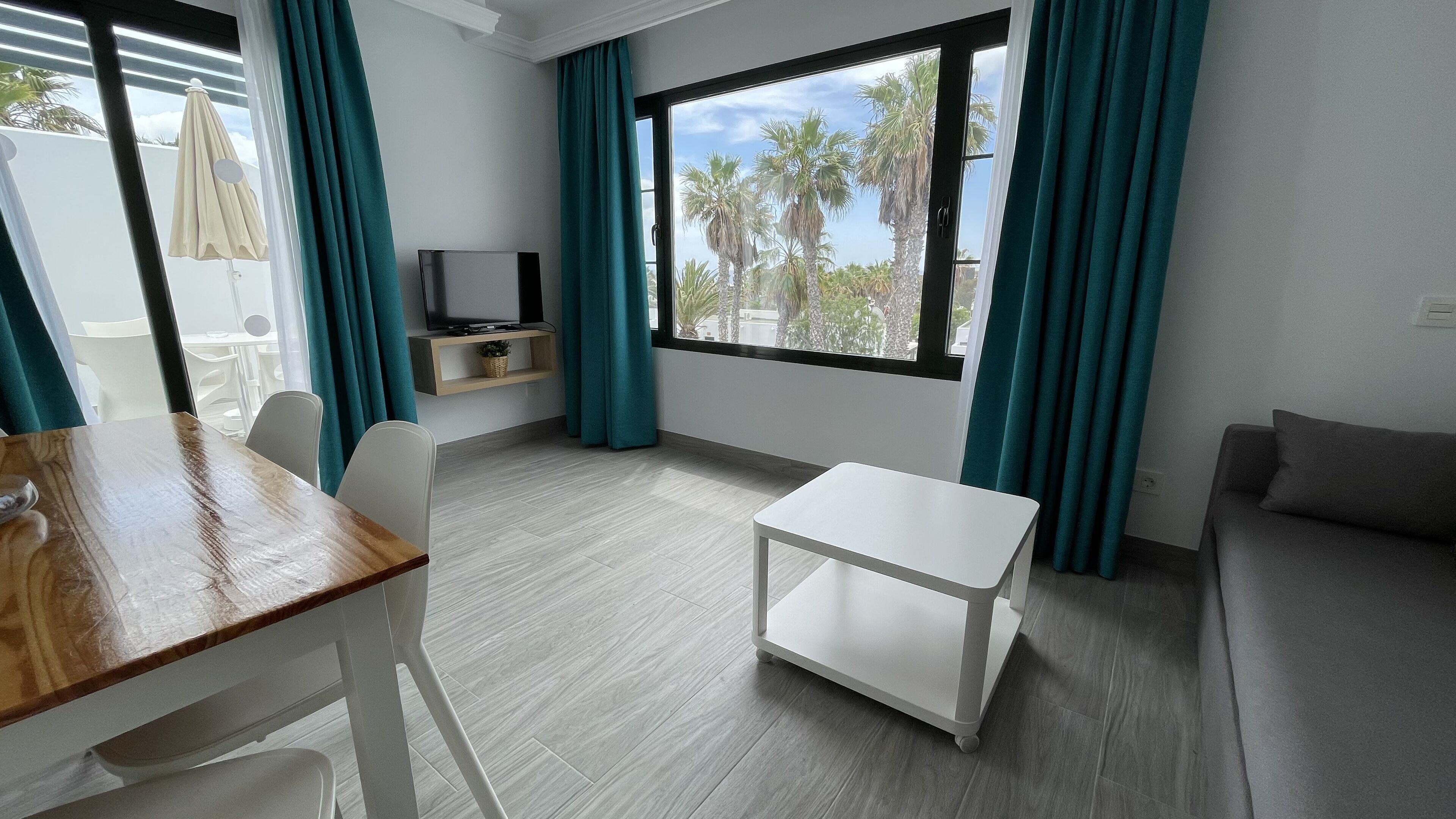 Een moderne woonkamer met een televisie, een eettafel, stoelen, een bank en uitzicht op palmbomen door grote ramen.