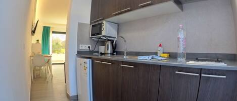 A modern kitchen with a microwave, kettle, and a sink. There is a small refrigerator and a dining area visible in the background.