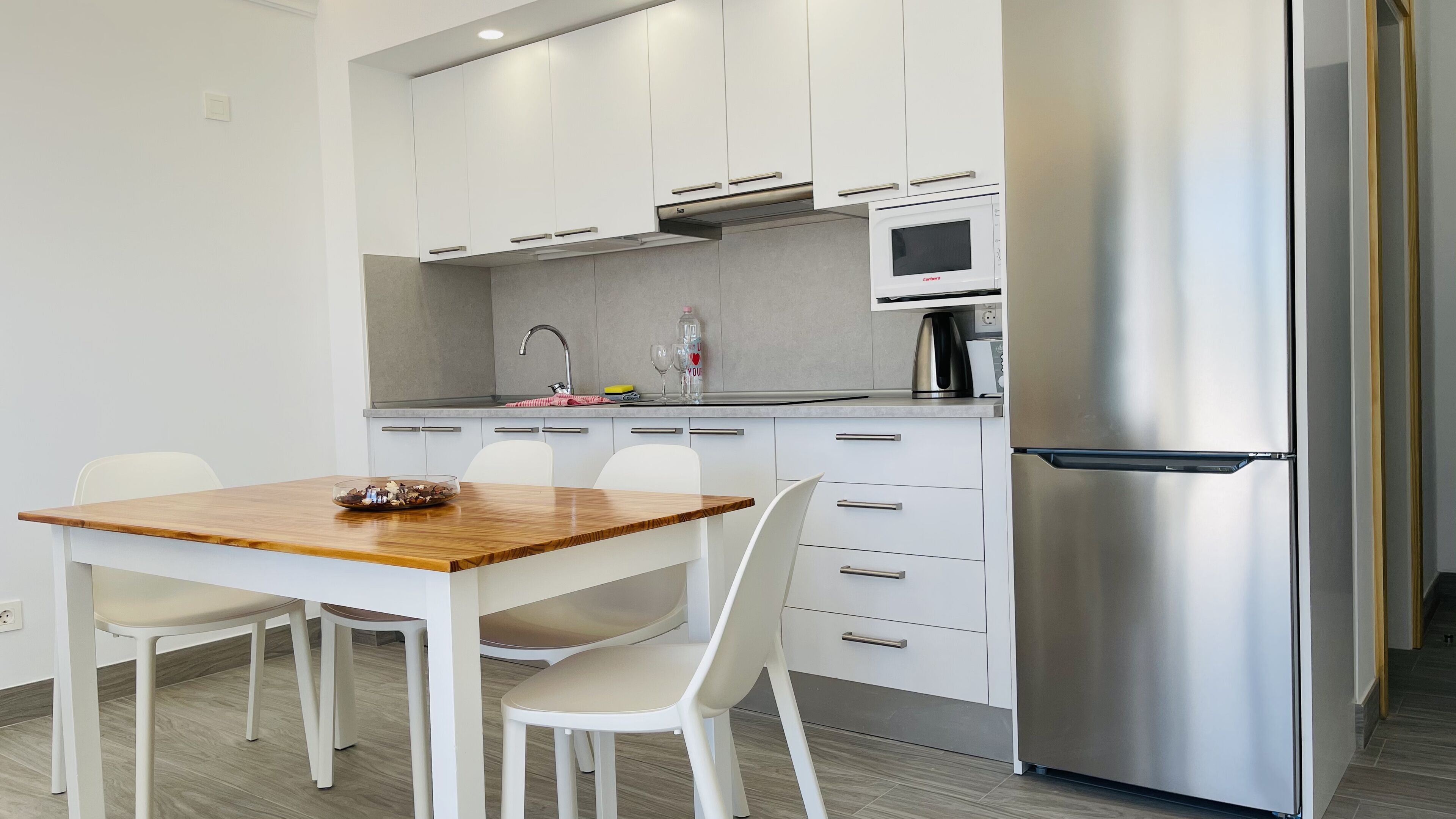 Una cocina moderna con armarios blancos, un refrigerador de acero inoxidable y una mesa de comedor de madera con sillas.