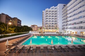 Une piscine entourée de chaises longues et de parasols, le tout adossé à un bâtiment moderne à plusieurs étages et doté de balcons.
