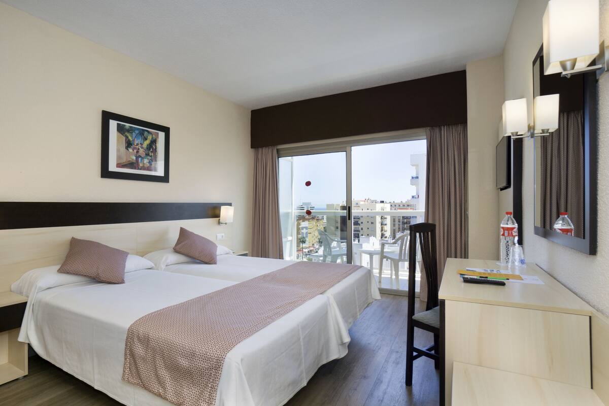 A hotel room with a large bed, a desk, a chair, a balcony with a view of the city, and a painting on the wall.