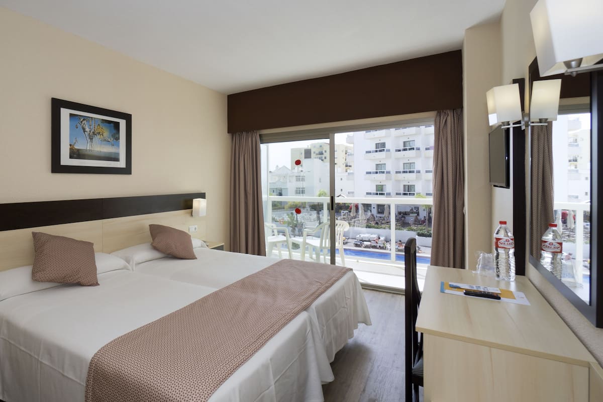A hotel room with a large bed, a bedside table, a desk with a chair, a window with curtains, and a view of buildings outside.
