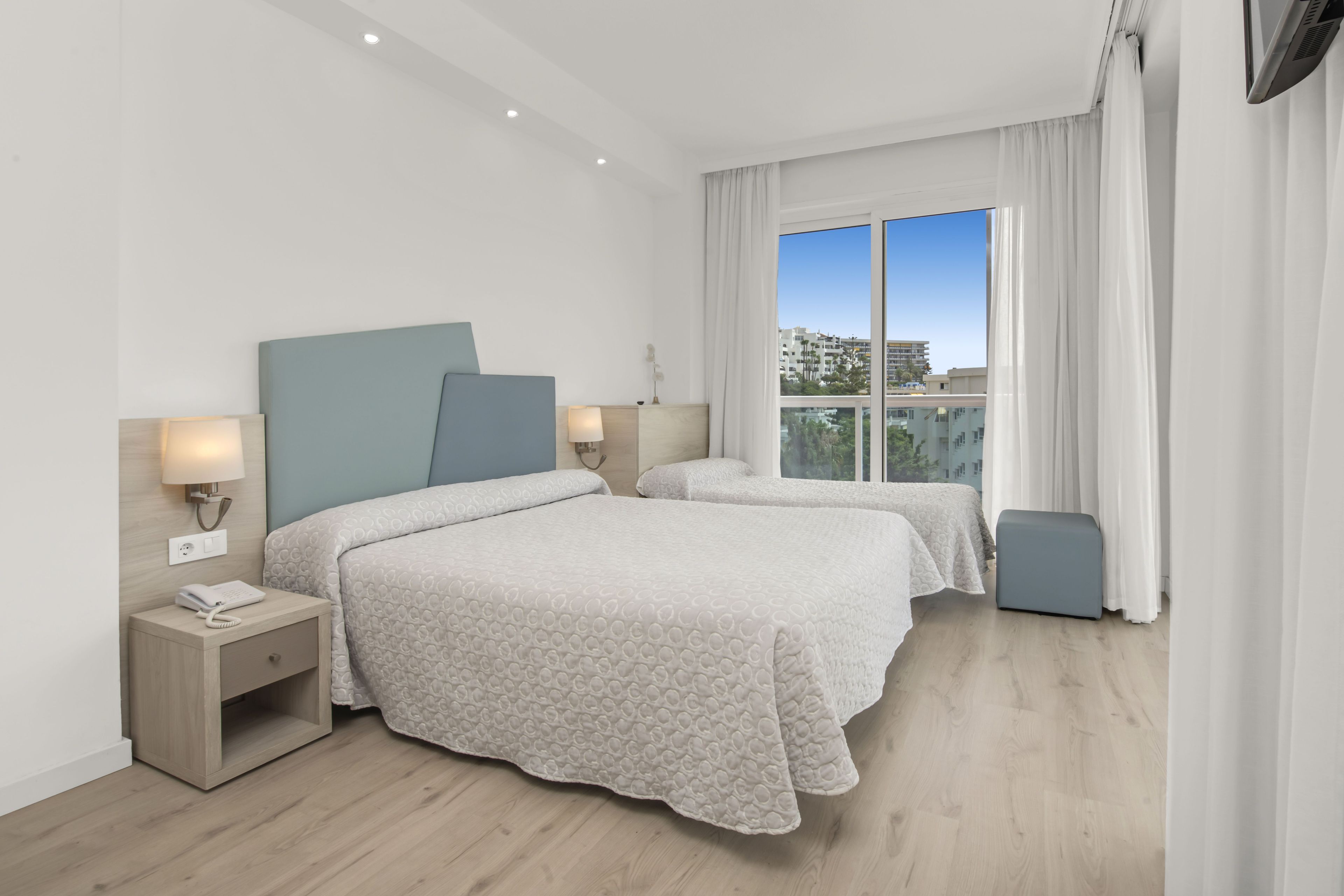A hotel room with two beds, a nightstand, a lamp, and a view of the city.