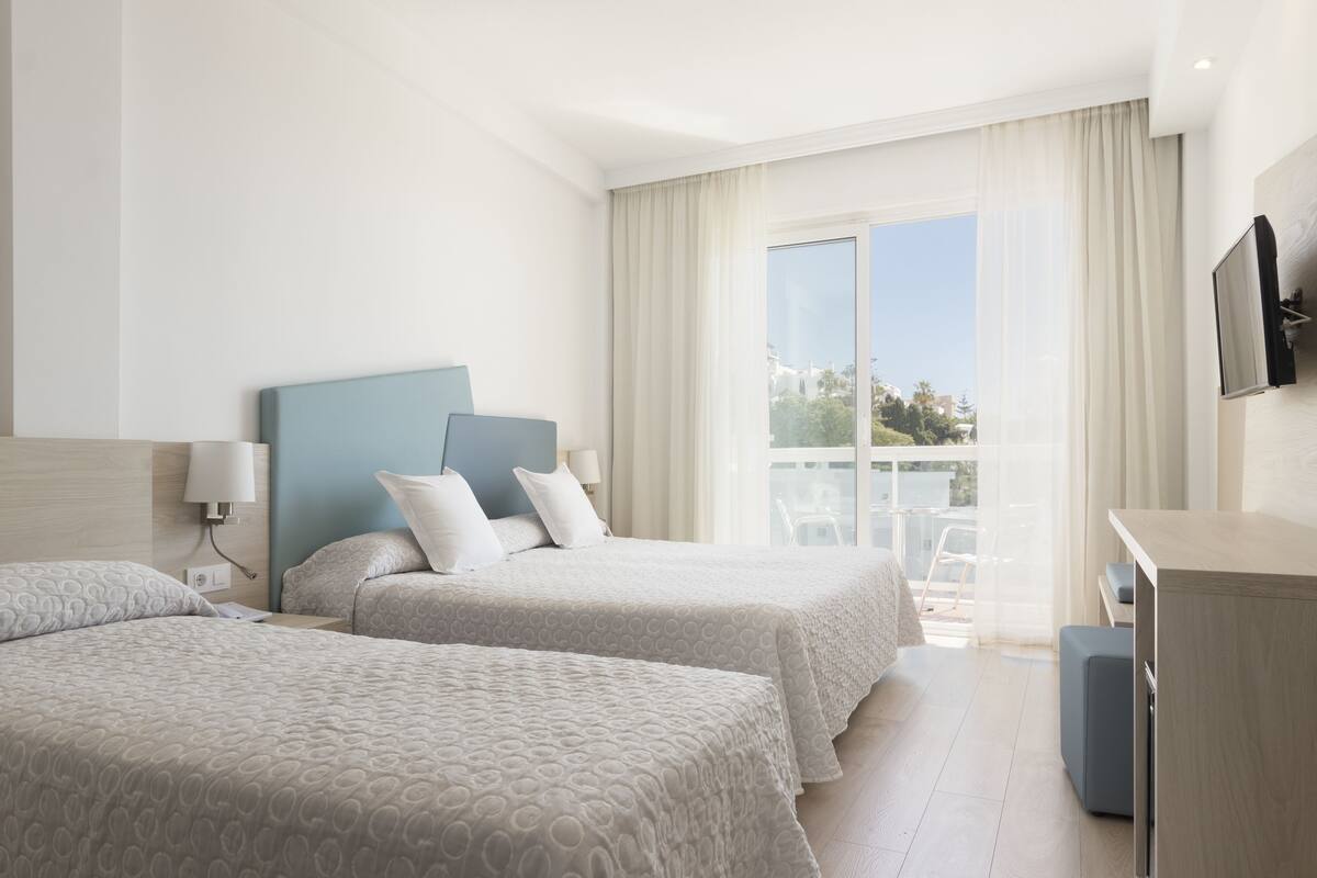 A hotel room with two beds, a TV mounted on the wall, and a balcony with a view.