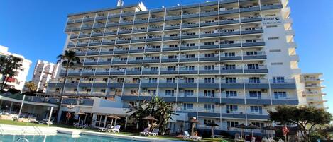 A multi-story hotel with balconies, a swimming pool, and a clear blue sky.