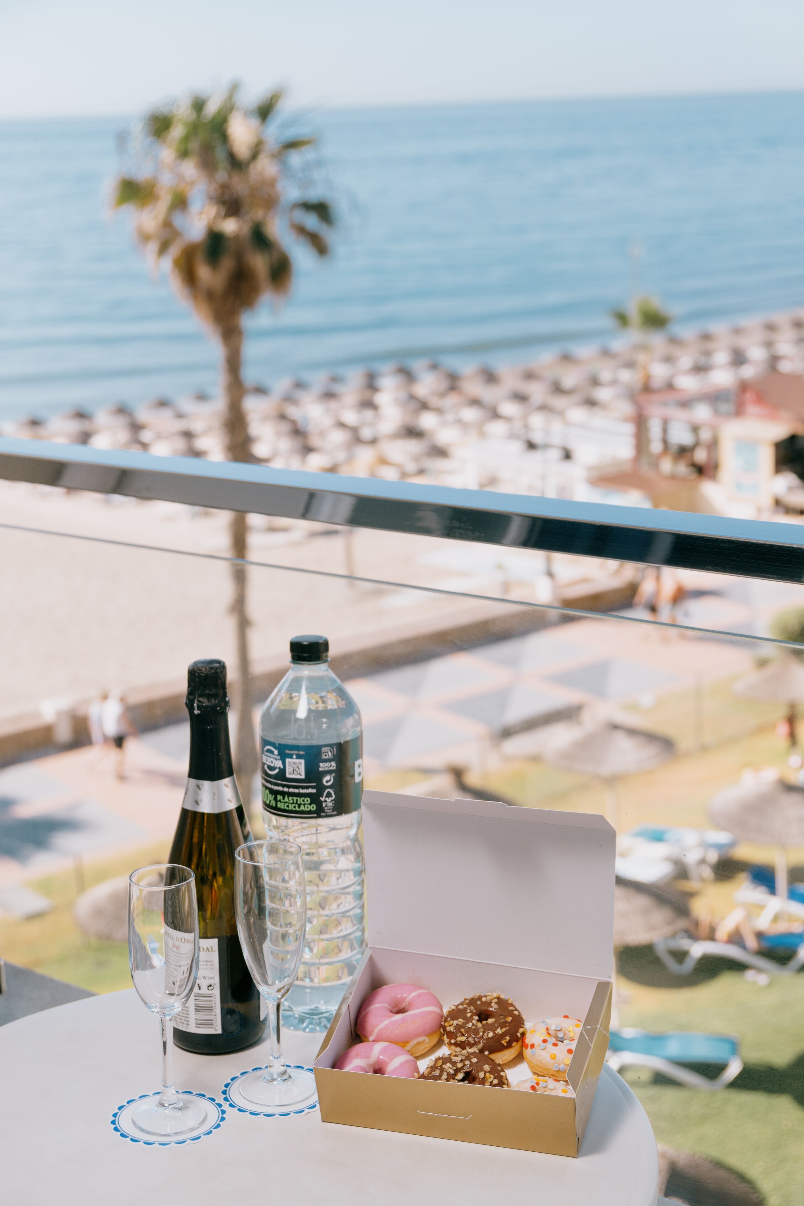 A table with a bottle of wine, two glasses, and a box of assorted donuts, overlooking a beach with lounge chairs and umbrellas.