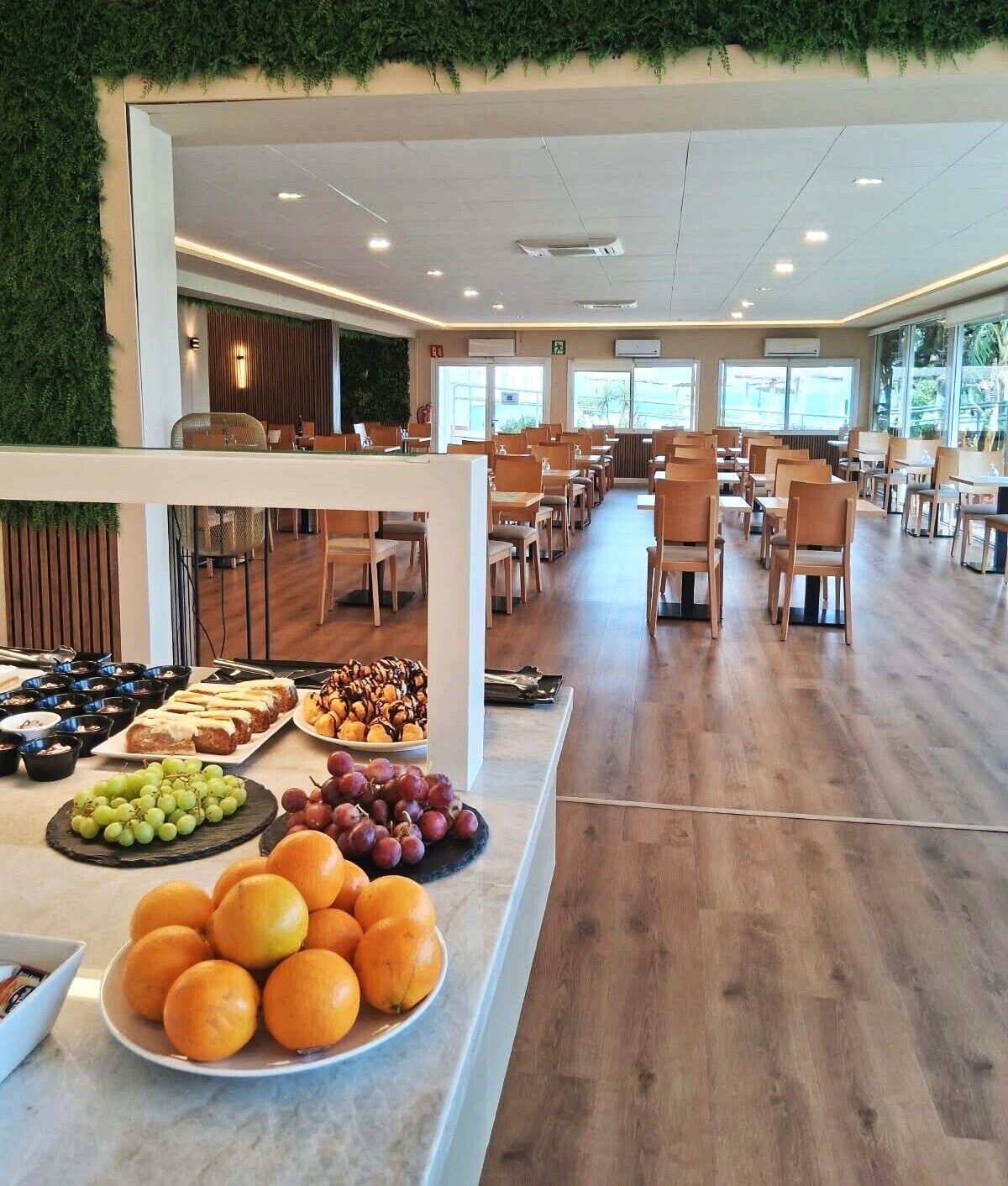 Une cafétéria spacieuse avec des tables et des chaises en bois, un buffet proposant des fruits et des pâtisseries, et de grandes fenêtres laissant entrer la lumière naturelle.