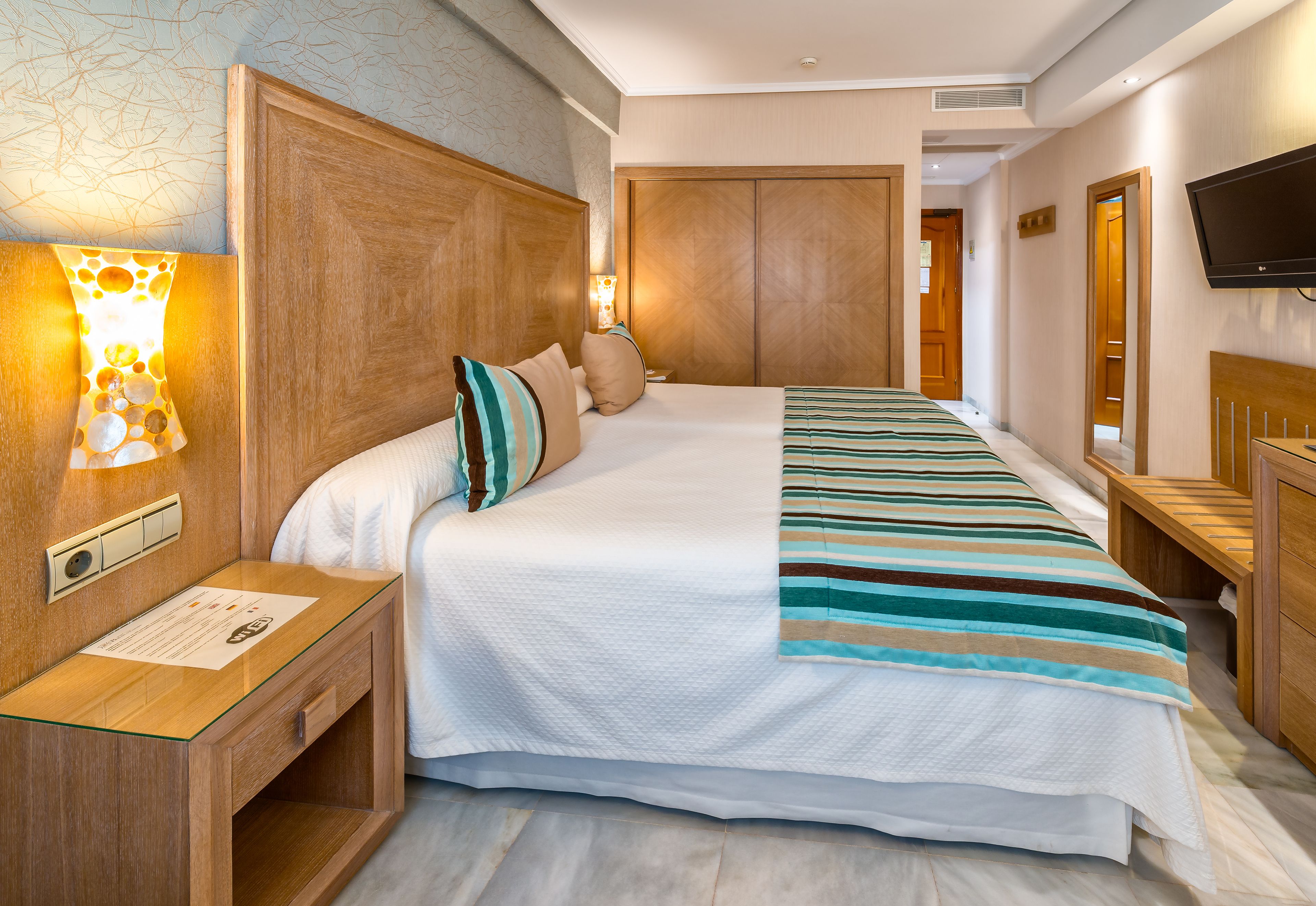 A hotel room with a large bed, wooden headboard, bedside tables, a flat-screen TV, and a wall-mounted lamp.