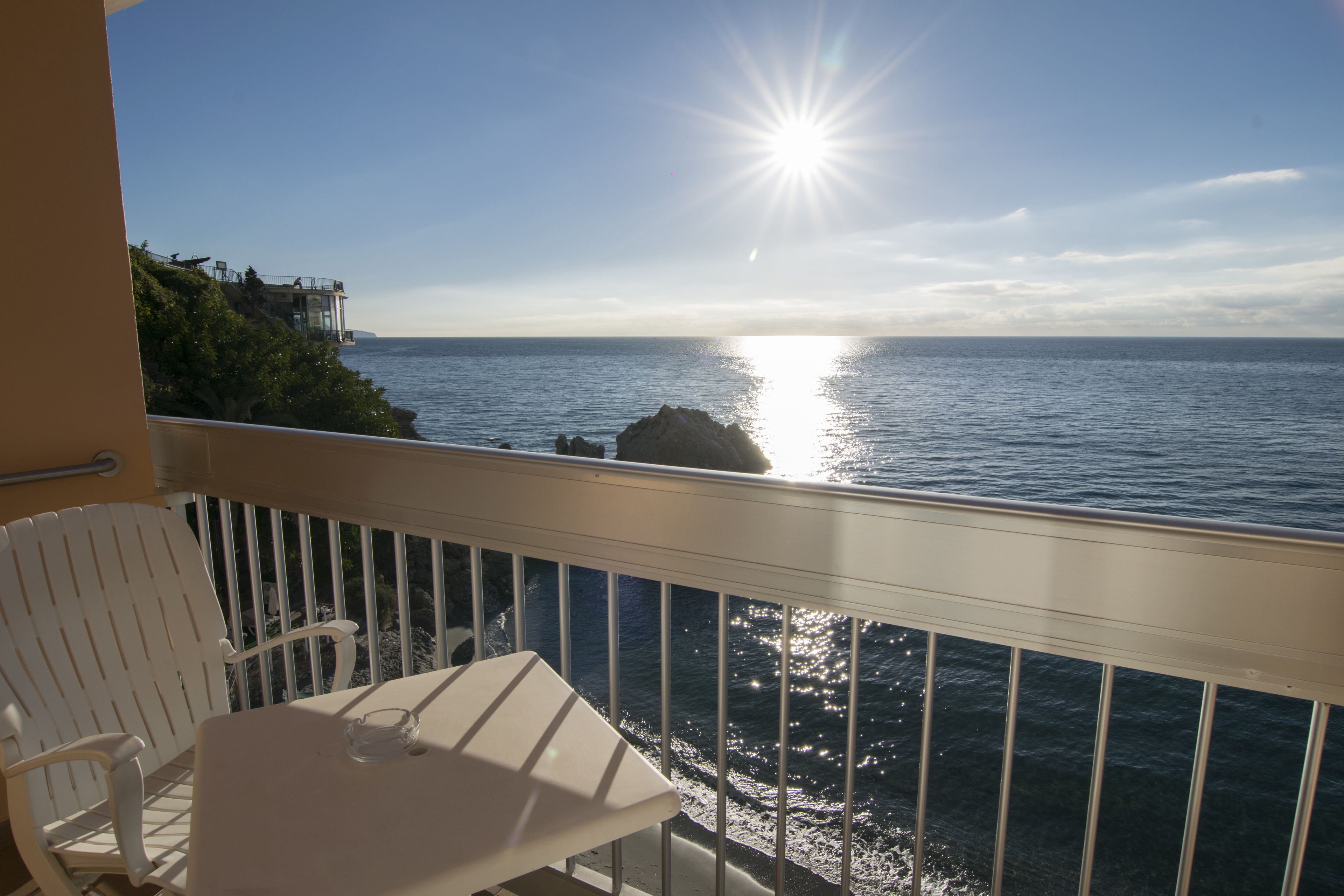 A balcony with a white chair and table, overlooking a coastal view with the sun reflecting on the water.