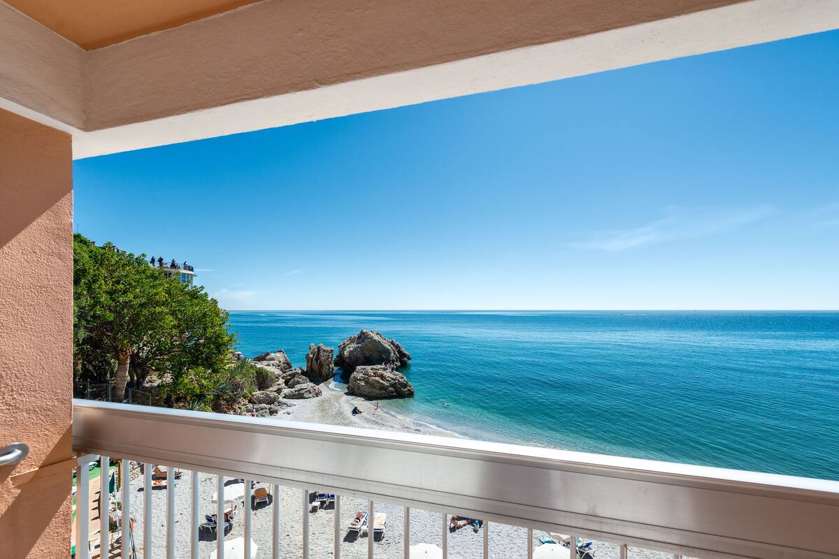 Een balkon met uitzicht op een strand, rotsformaties en helderblauw water.