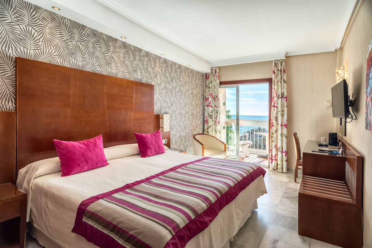 Een hotelkamer met een groot bed, een houten hoofdeinde, uitzicht op zee, een bureau en een televisie.