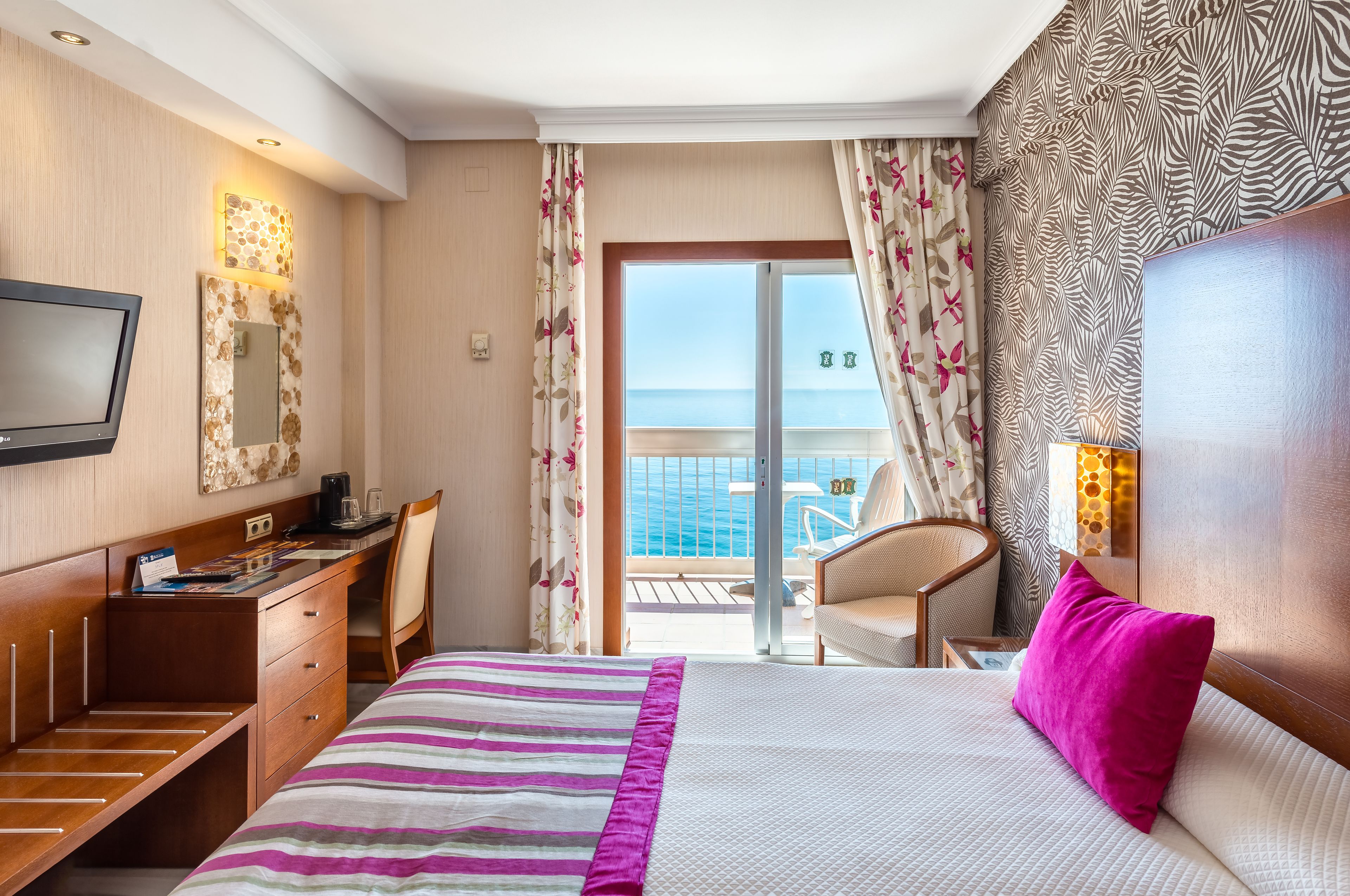 Ein Hotelzimmer mit Bett, Schreibtisch, Stuhl, Fernseher und einem Balkon mit Meerblick.